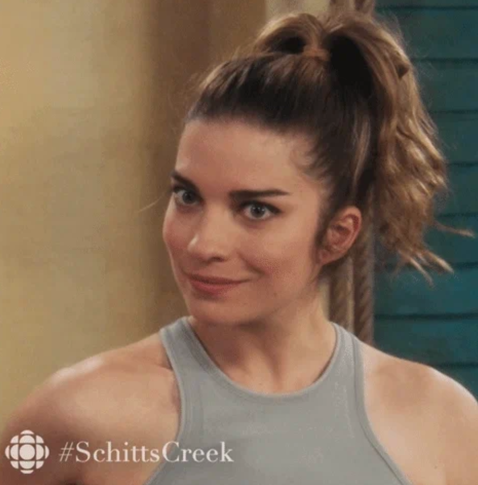 A person with a high ponytail smiles in a scene from "Schitt's Creek." They wear a sleeveless top. #SchittsCreek is visible in the image
