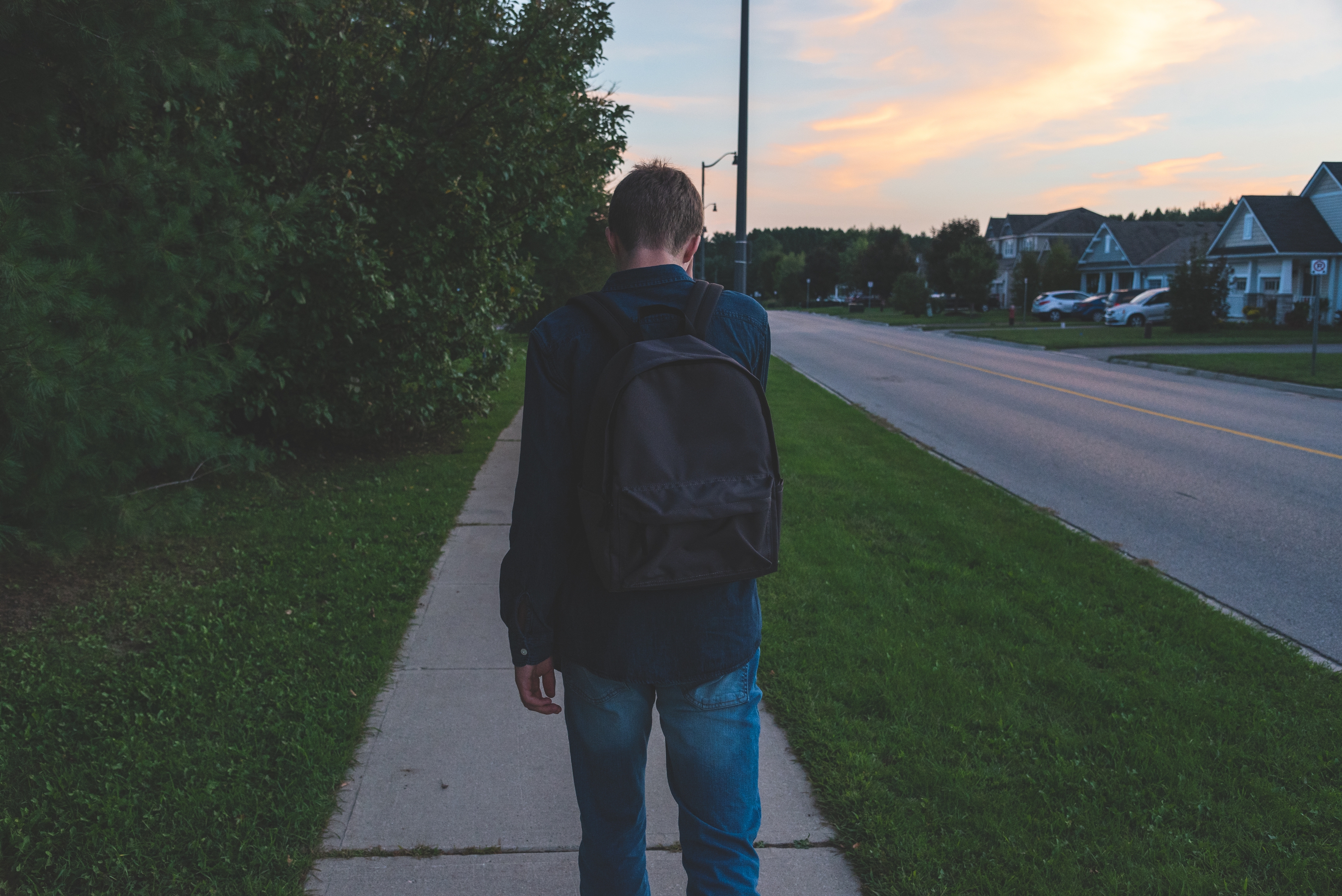 Person walking on sidewalk with a backpack, heading towards a residential area at dusk