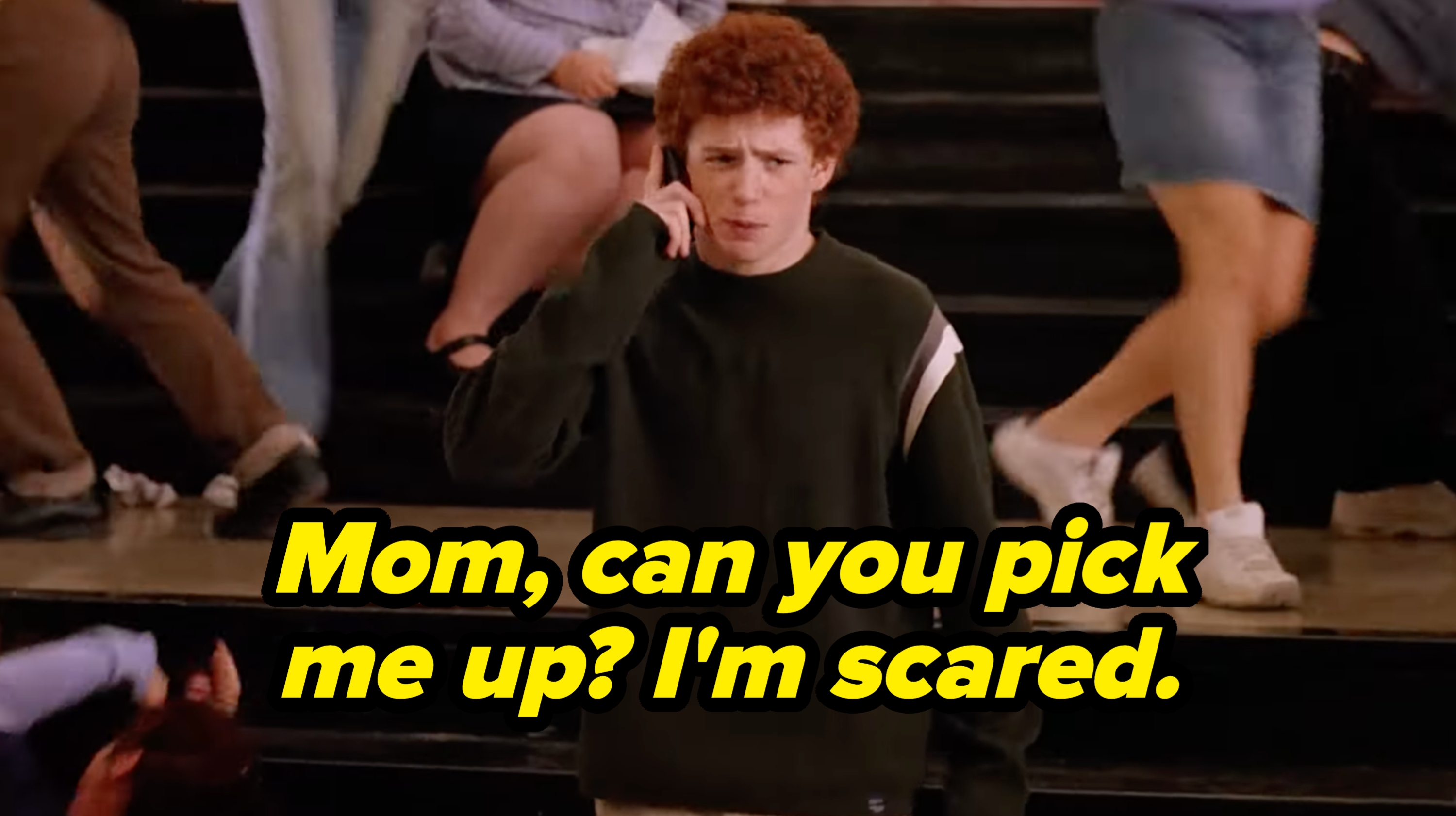 Person with curly hair stands on a staircase, appearing worried while talking on a cellphone, surrounded by people moving past