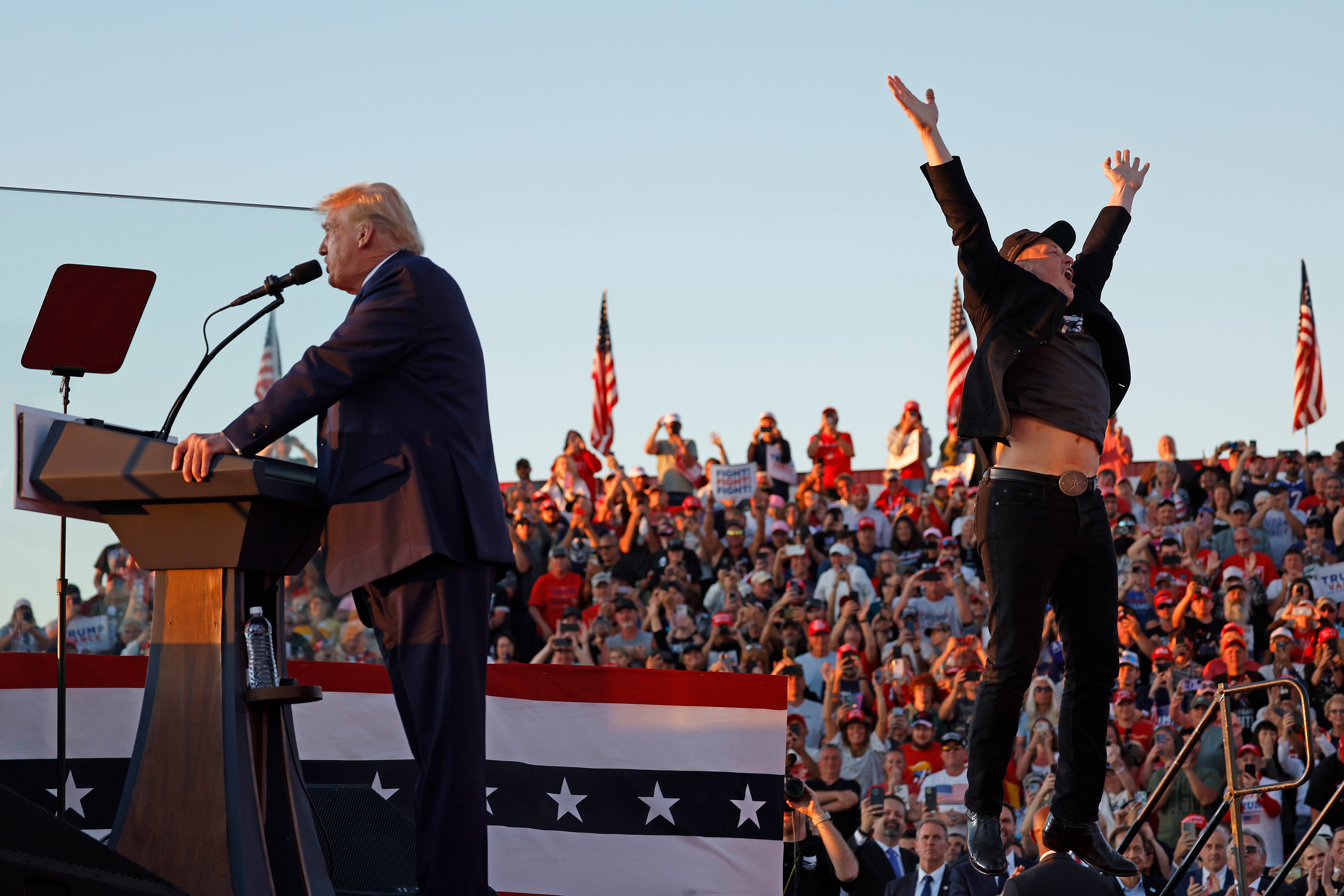 A person in a suit speaks at a podium with a large crowd in the background. Another person nearby raises their arms energetically