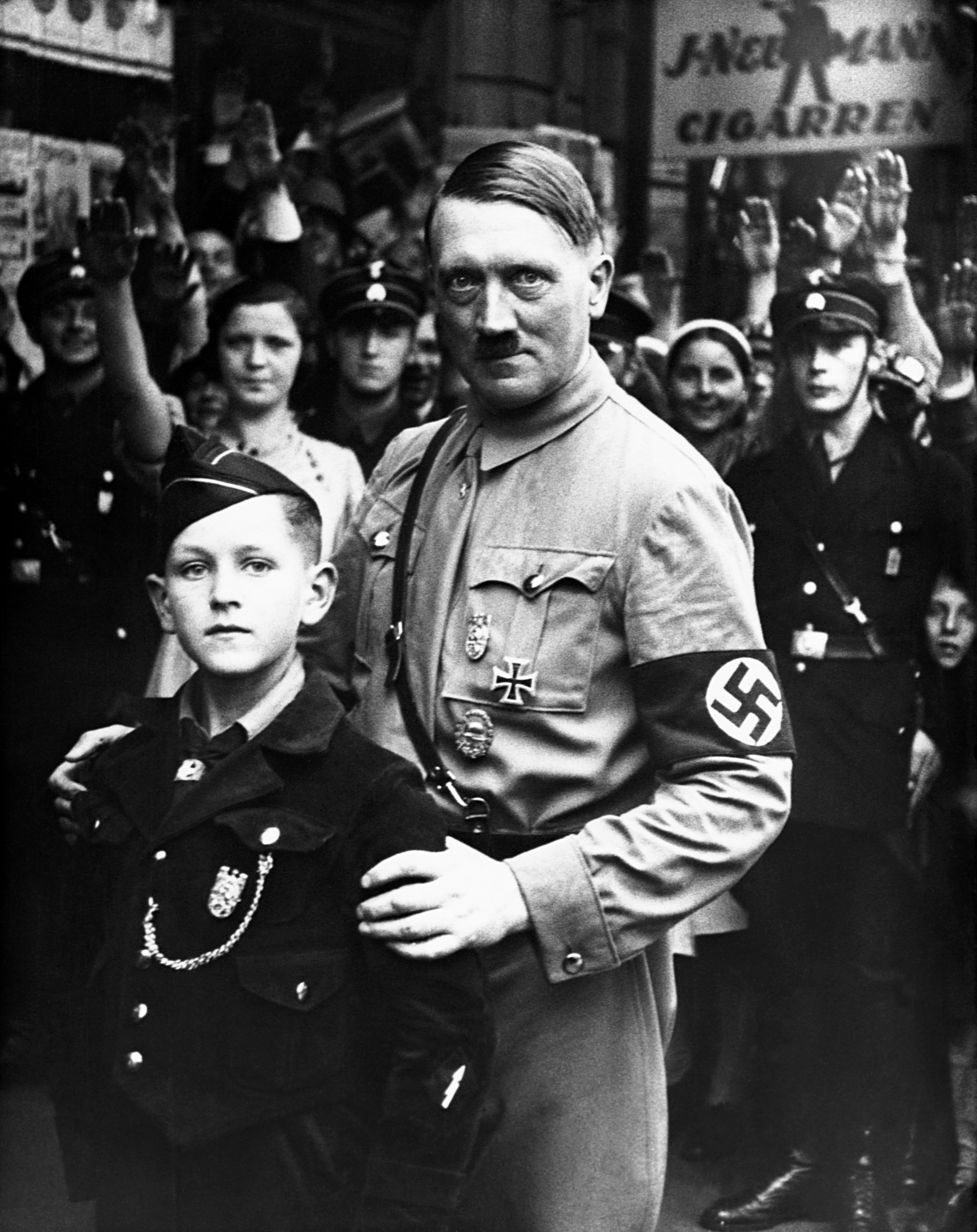 A man in a uniform with a swastika armband stands next to a child also in uniform, amid a group of saluting people