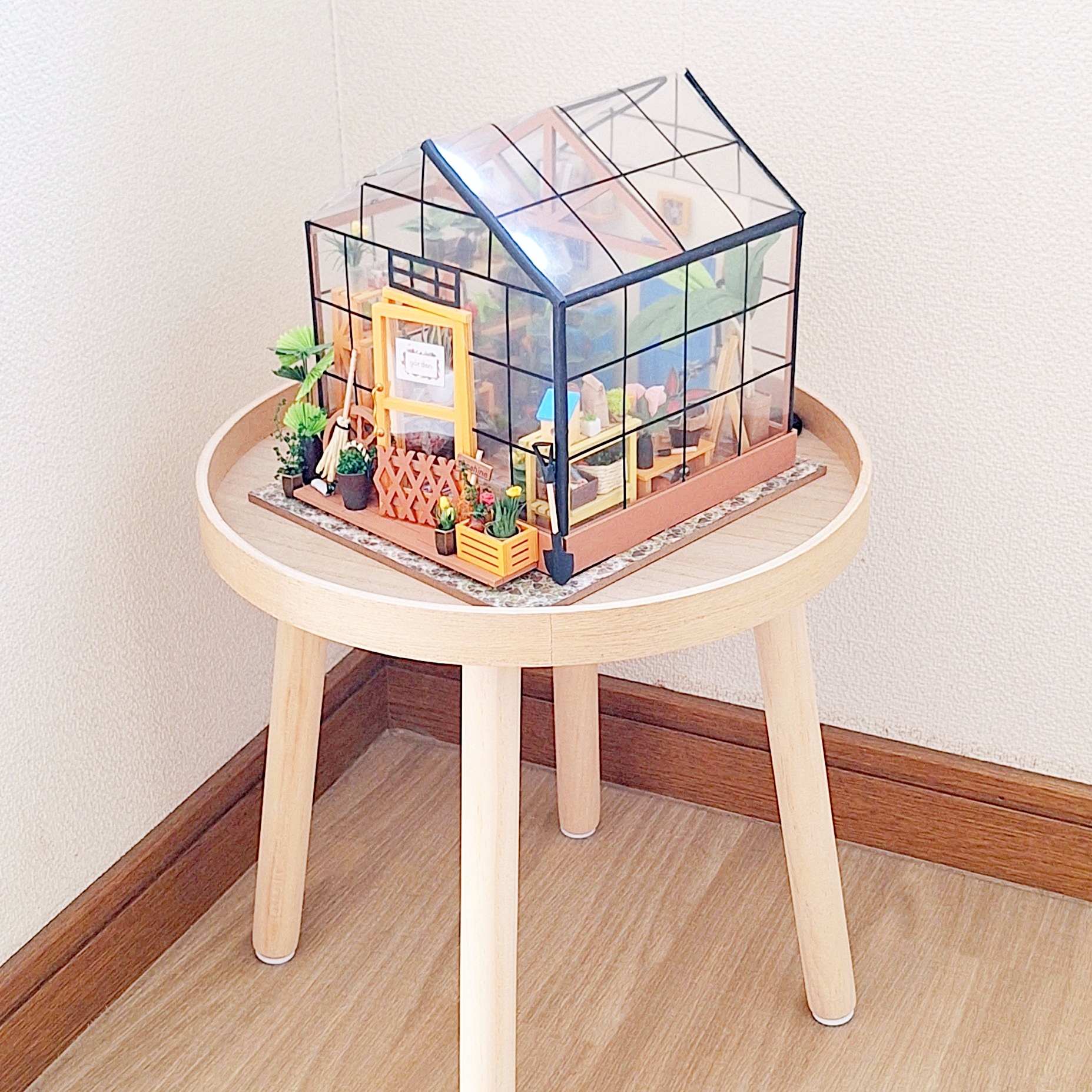 小さな木製の棚の上に置かれた、ミニチュアのガラス温室模型。植物や小物が細かく配置されている。