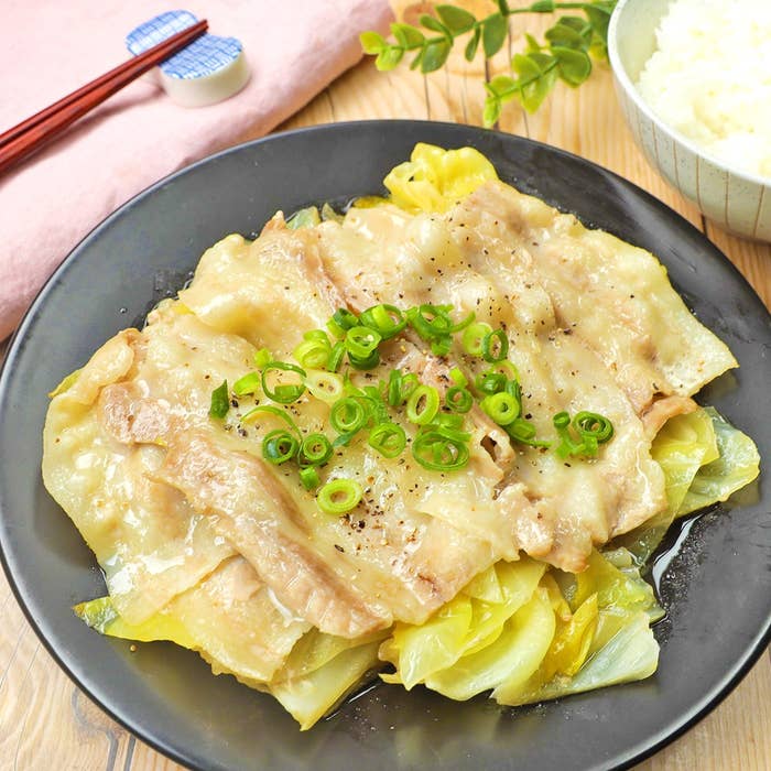 野菜と豚肉の蒸し料理が黒い皿に盛られ、ネギが上に飾られた料理。横にご飯と割り箸が添えられています。