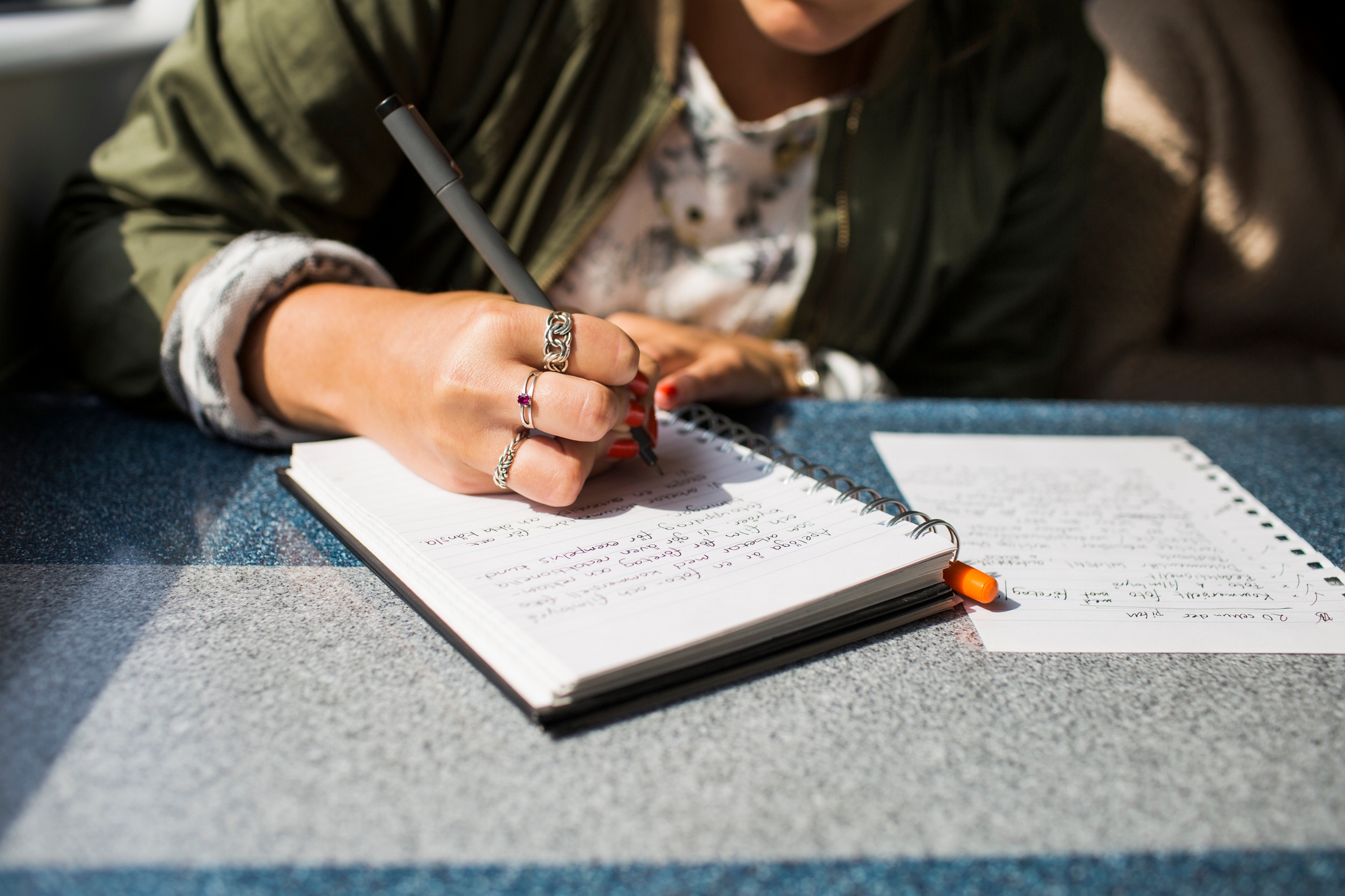 Person writing in a notebook at a table, with a page of handwritten notes beside them. Wearing a jacket and silver rings