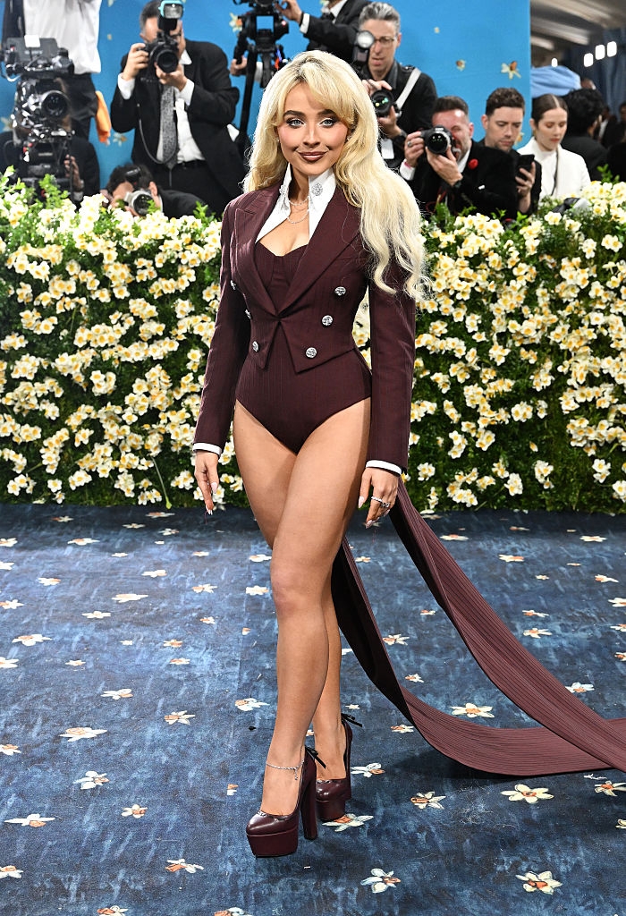 A person on the red carpet wears a burgundy blazer-style bodysuit with a long train, paired with matching high heels