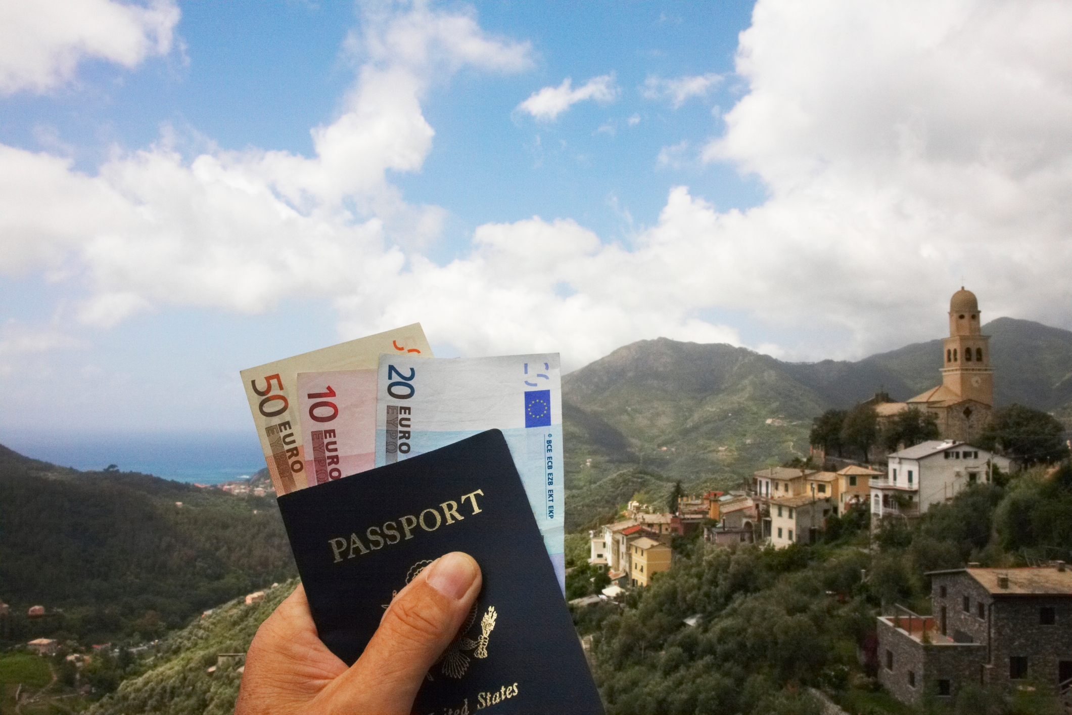 Hand holding a passport and Euro notes against a scenic village backdrop with rolling hills and a tower