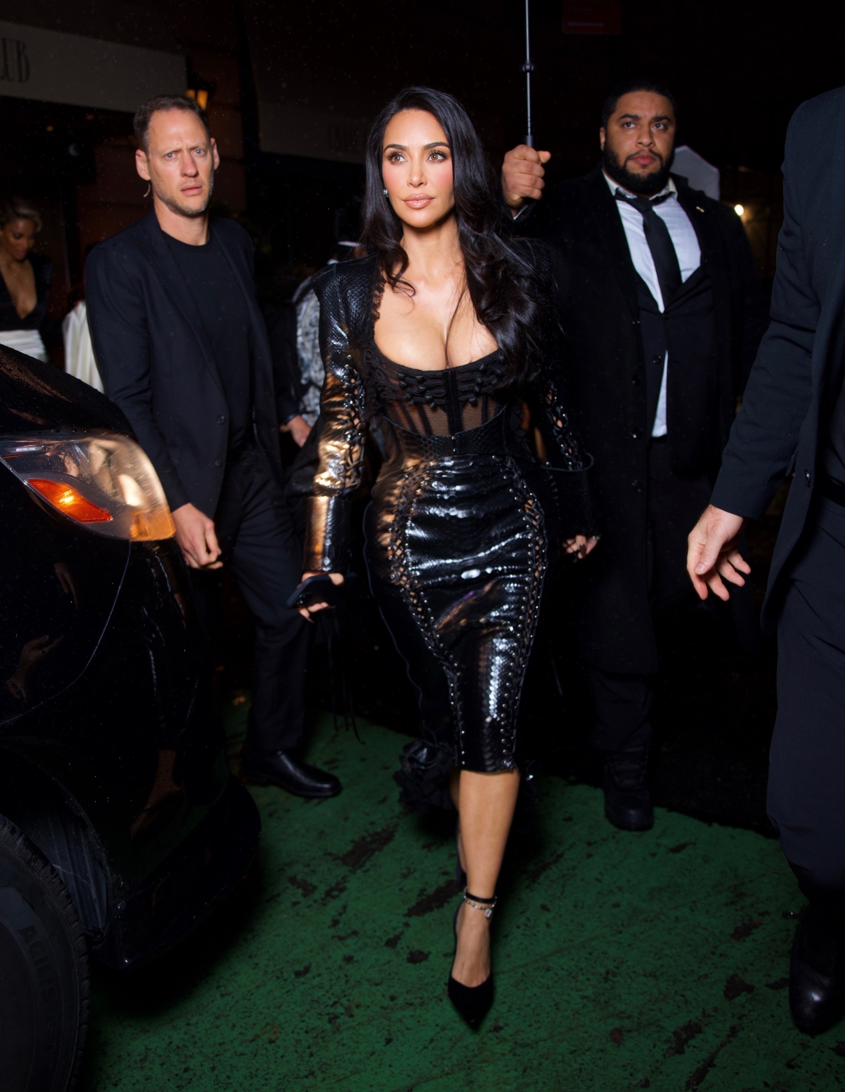 Woman in a fitted, glossy black outfit walks on a street, surrounded by security personnel