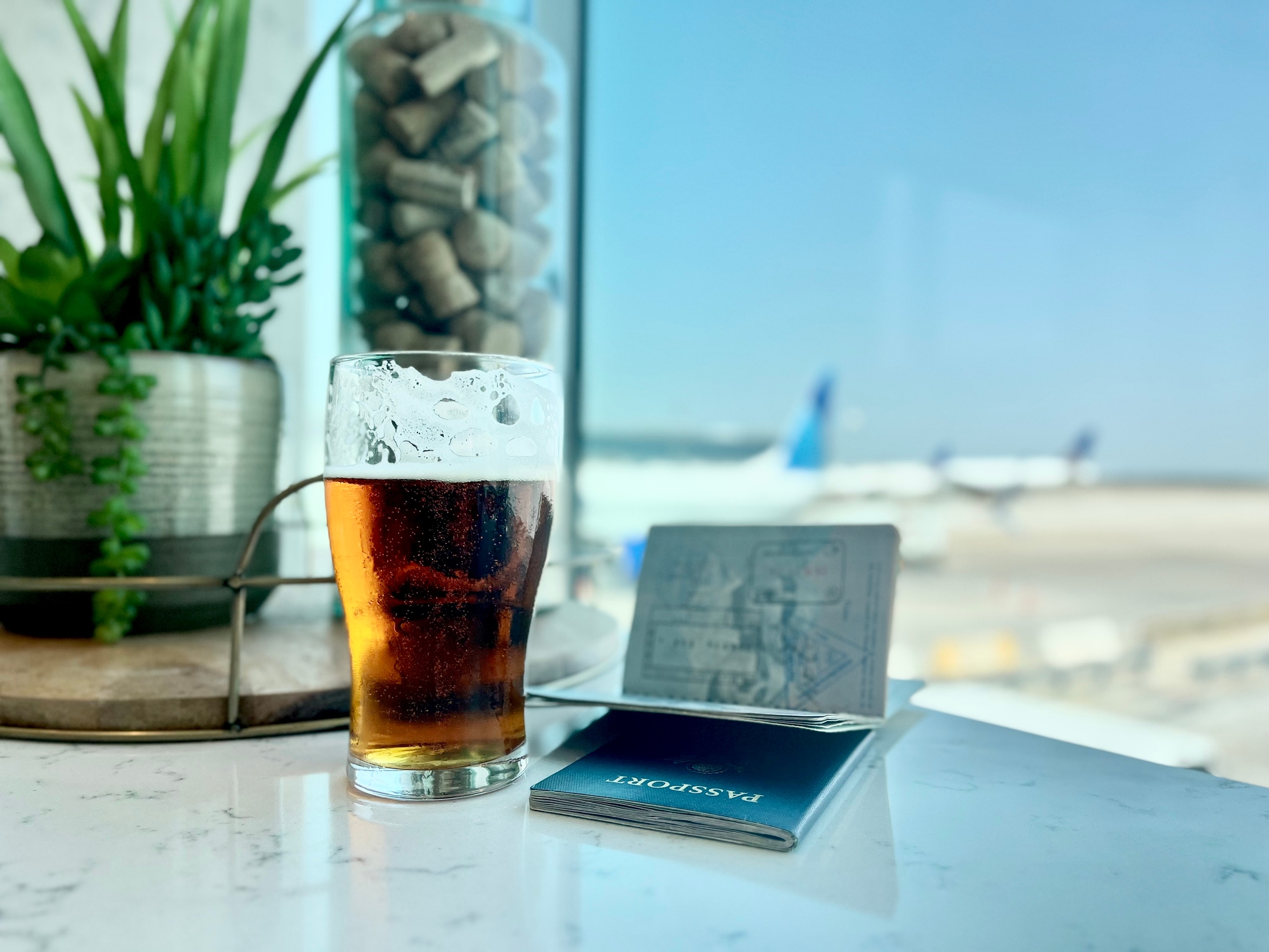 A passport and a pint of beer are on a table at an airport lounge with a blurred view of airplanes outside