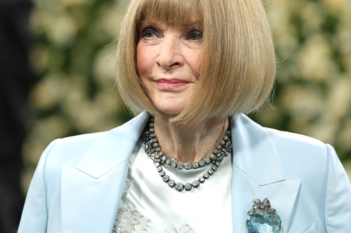 Person in elegant attire with a blazer, intricate necklace, and heart-shaped brooch, standing in front of a floral backdrop