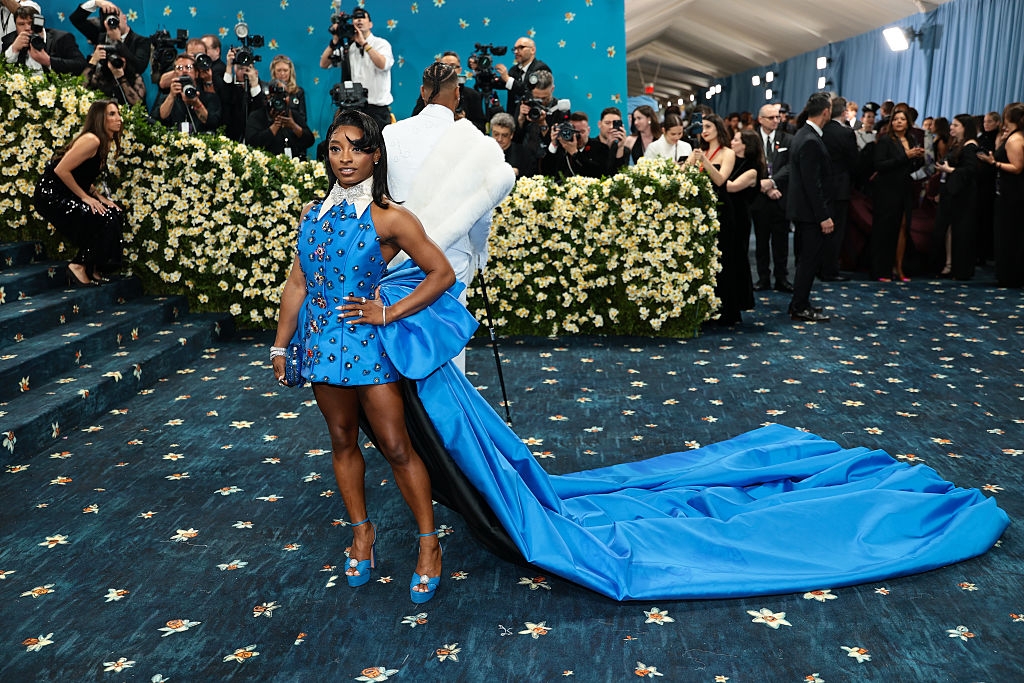 A woman poses on the red carpet in a stylish blue dress with a long train. Photographers capture the moment in the background