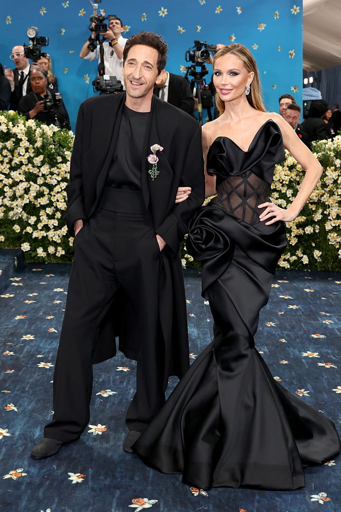Two individuals pose on a red carpet. One wears a black suit with a floral detail; the other wears a black gown with mesh and floral accents