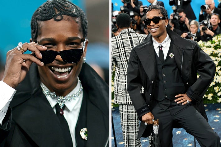 Person in stylish tailored suit on a red carpet; wearing sunglasses, layered necklaces, and rings, striking a confident pose