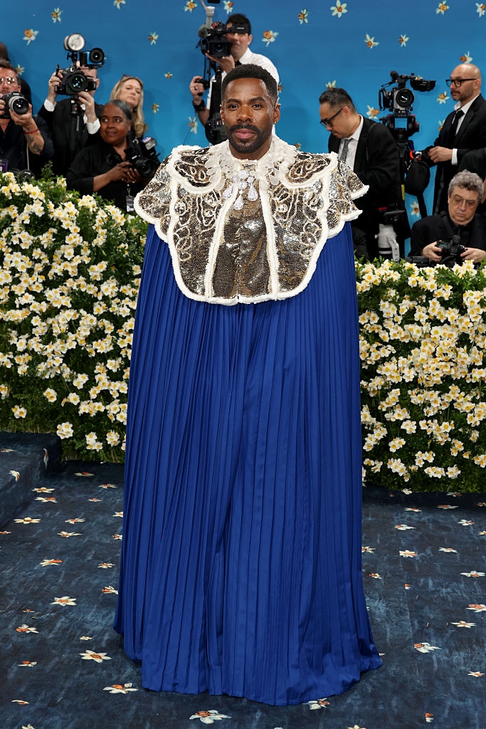 Person in elaborate cape with ornate collar poses on a floral-themed red carpet, surrounded by photographers