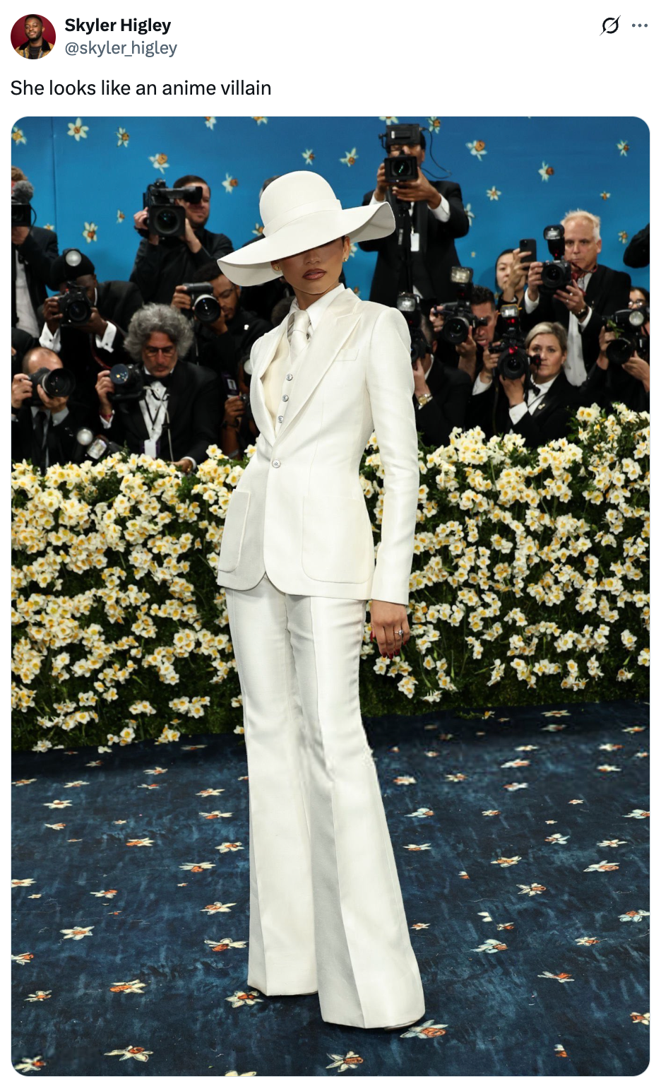 Person in a stylish, tailored white suit and wide-brimmed hat on a blue carpet, surrounded by photographers amidst floral decor