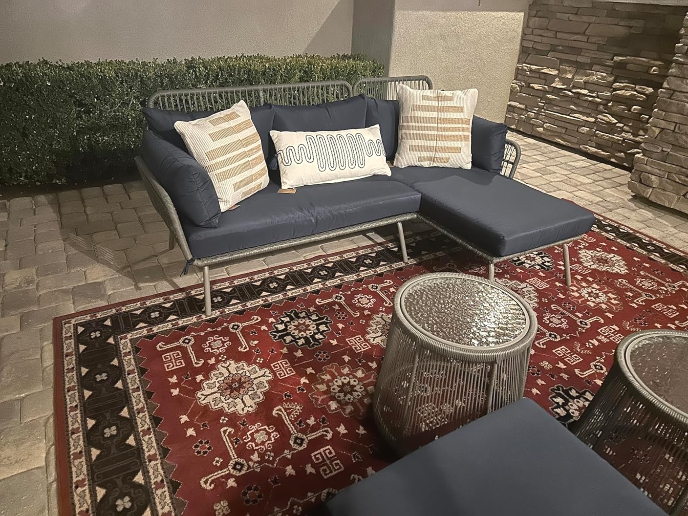 Outdoor seating area with a dark sofa, patterned pillows, and metallic tables on a decorative rug, set against stone and greenery
