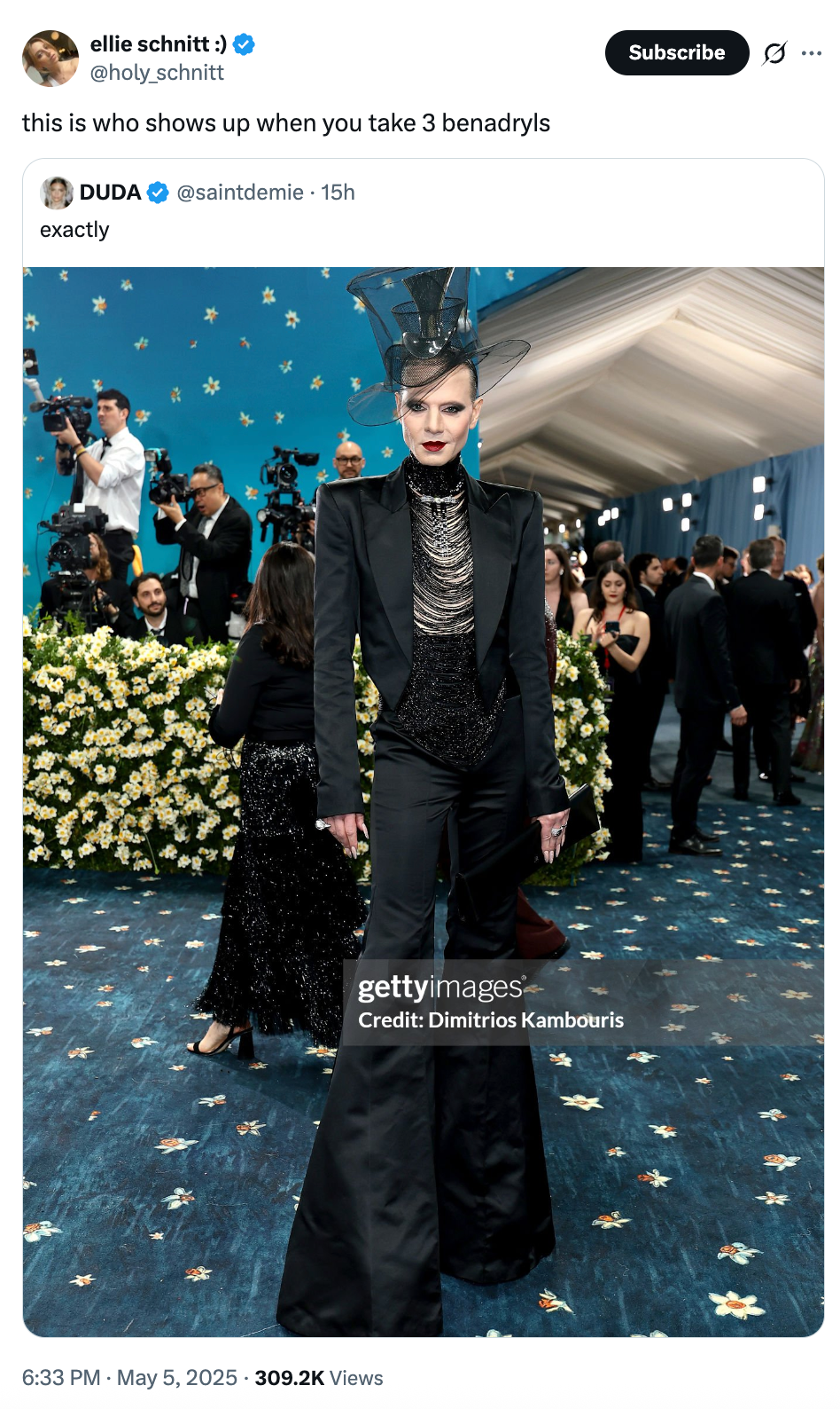 A person in an avant-garde black suit and intricate headpiece poses on a crowded event floor