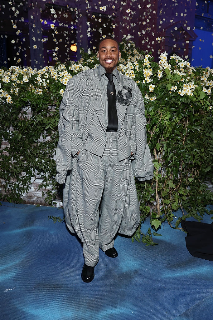 Person in oversized checkered suit with floral brooch, standing on a patterned carpet in front of greenery and flower backdrop