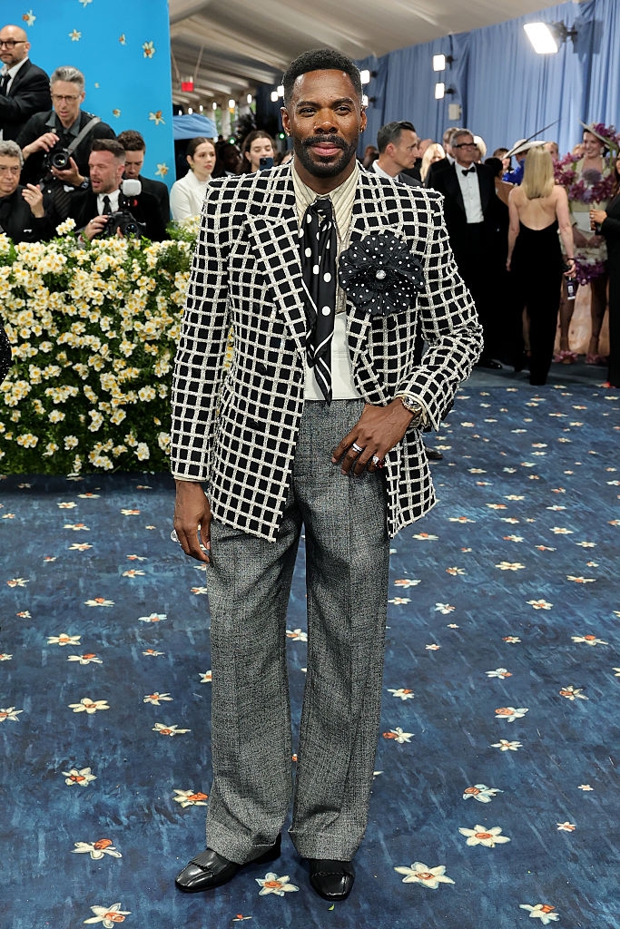 Man in a checkered suit with floral lapel and polka dot tie poses elegantly on a floral carpeted event