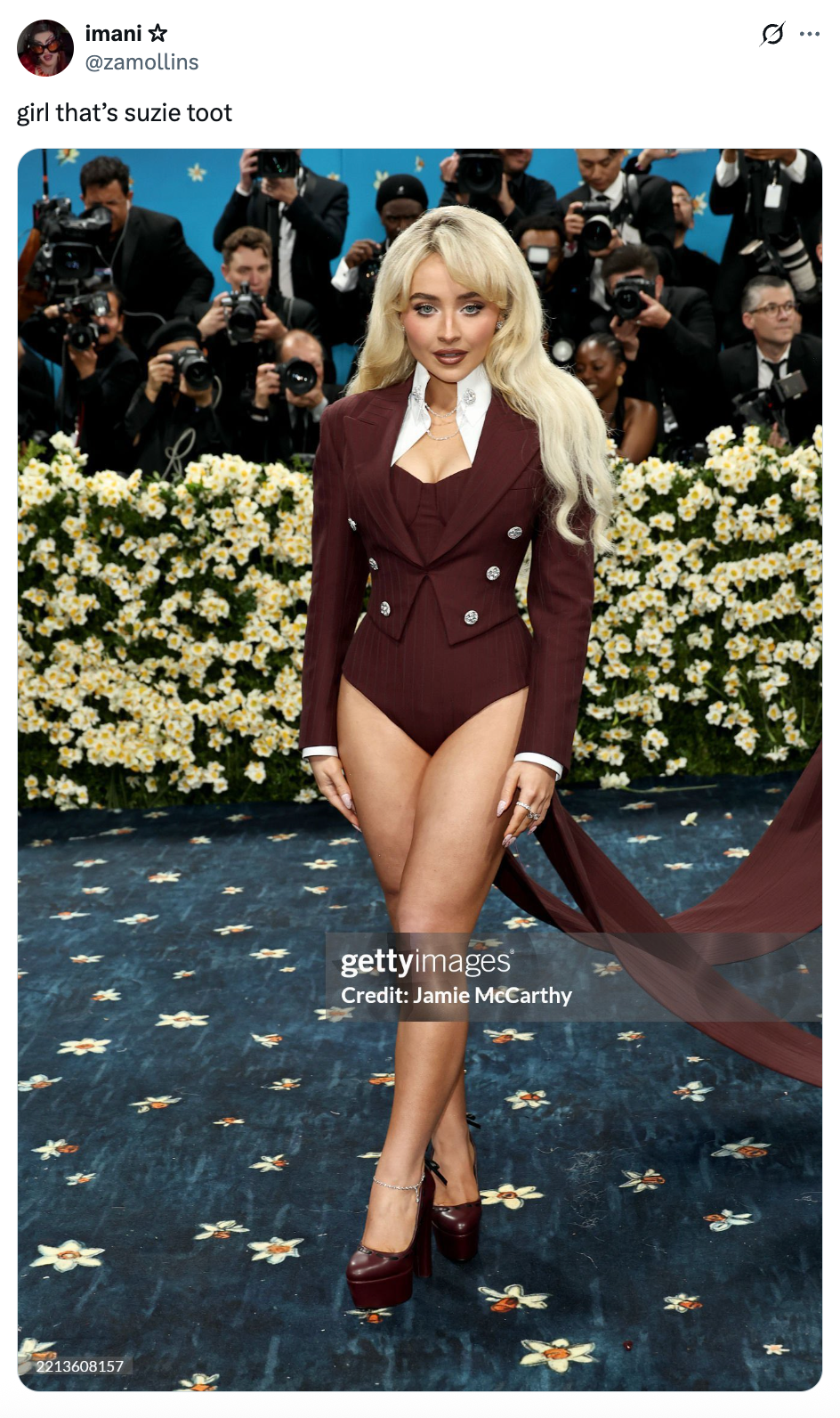 Celebrity wears a burgundy blazer-style bodysuit with a long train, posing in front of a floral backdrop at a red carpet event