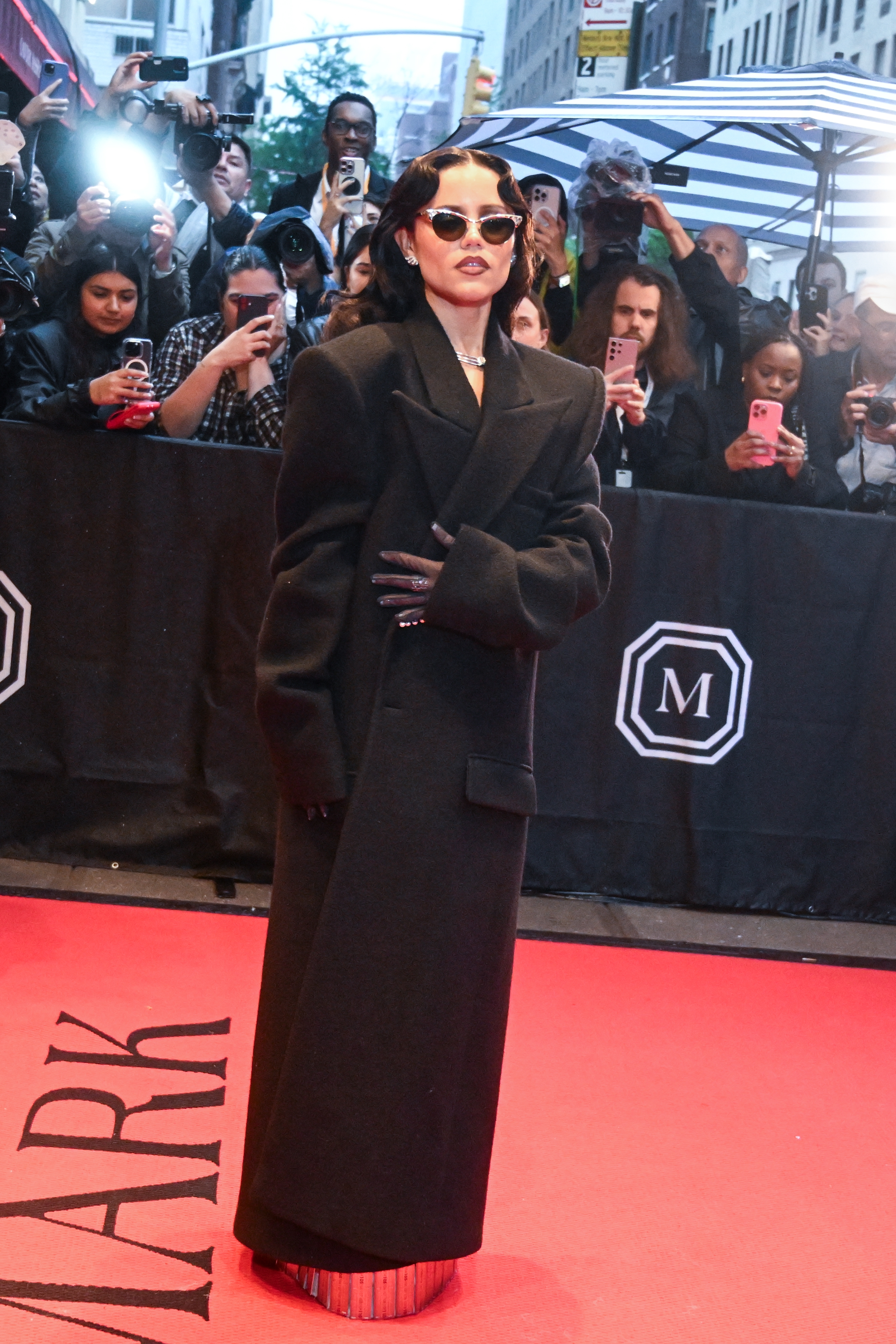 Jenna Ortega poses on red carpet in a stylish, oversized black coat and sunglasses, surrounded by photographers and fans