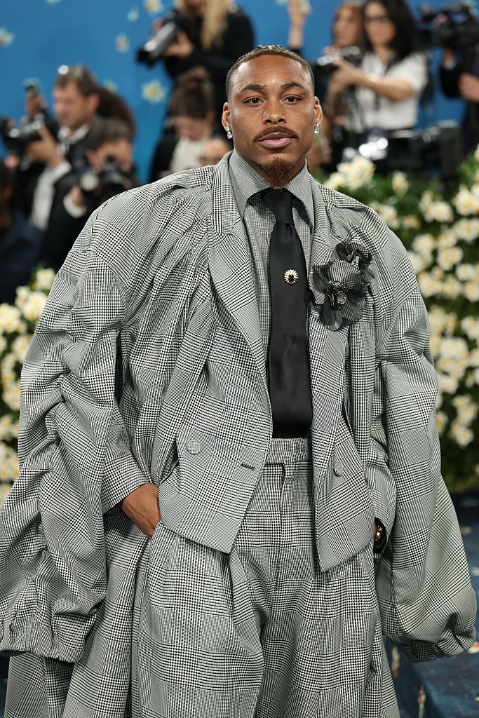 A person poses on the red carpet wearing an oversized plaid suit, a black tie, and a floral accessory, with photographers in the background