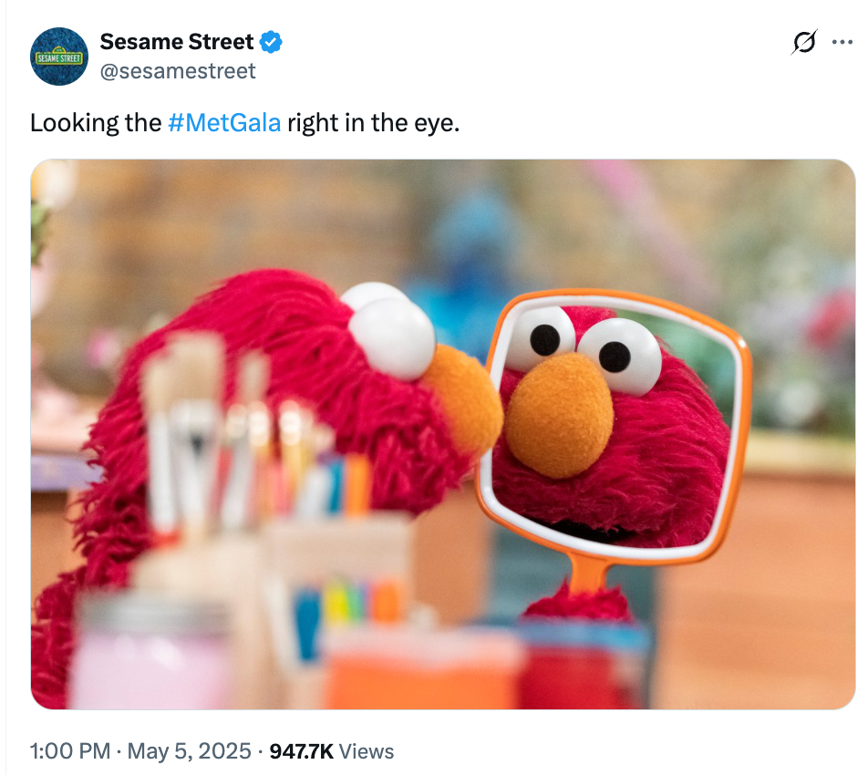 Elmo looks into a handheld mirror, surrounded by art supplies, in a playful nod to the Met Gala