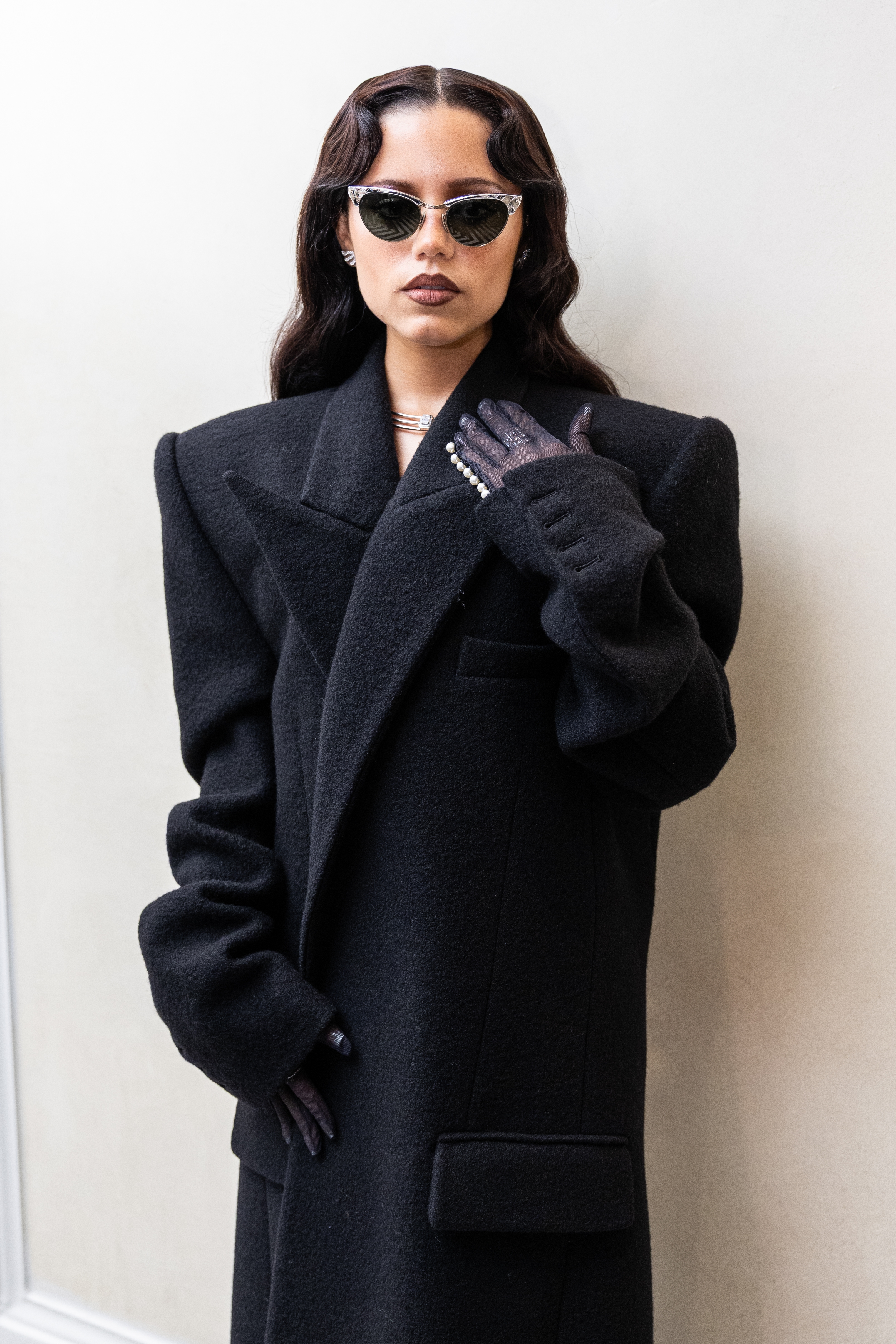 Jenna Ortega wearing a stylish, oversized coat and sunglasses with wavy hair, posing against a plain background