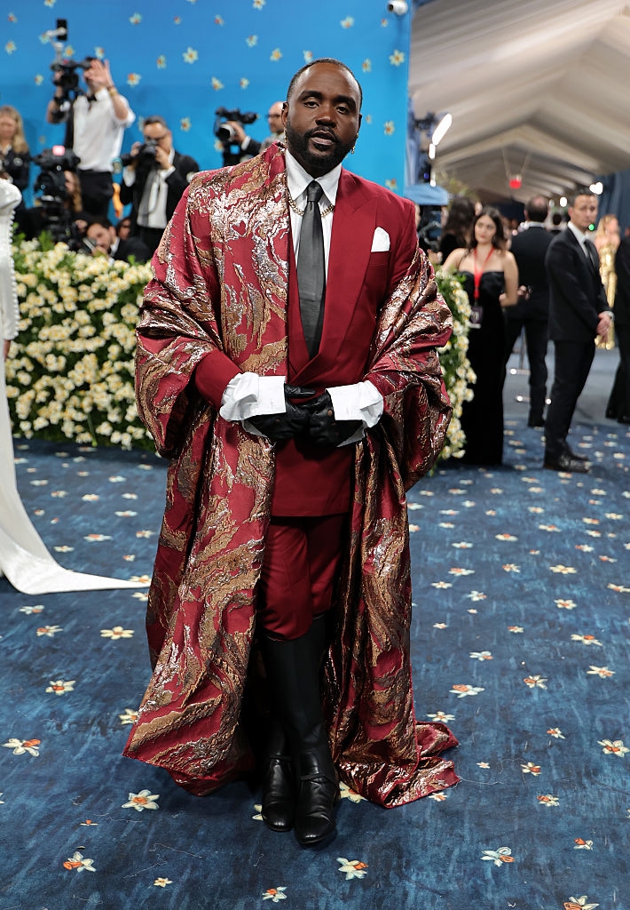 A celebrity in an ornate suit with a paisley-patterned cape, white shirt, and black tie on a blue carpet at a formal event