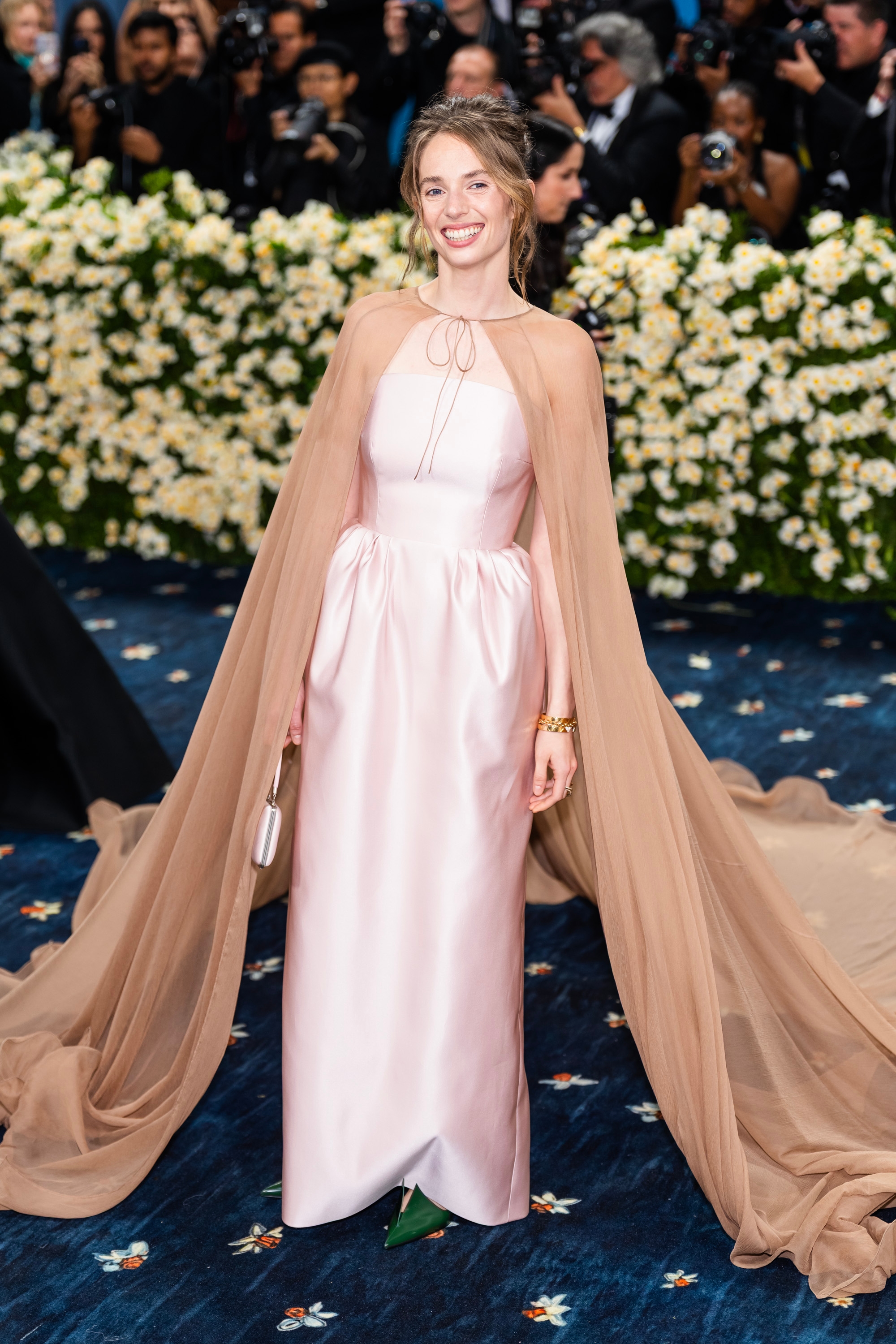 Individual in an elegant, sleeveless gown with a flowing cape poses on a red carpet, surrounded by photographers and floral backdrop