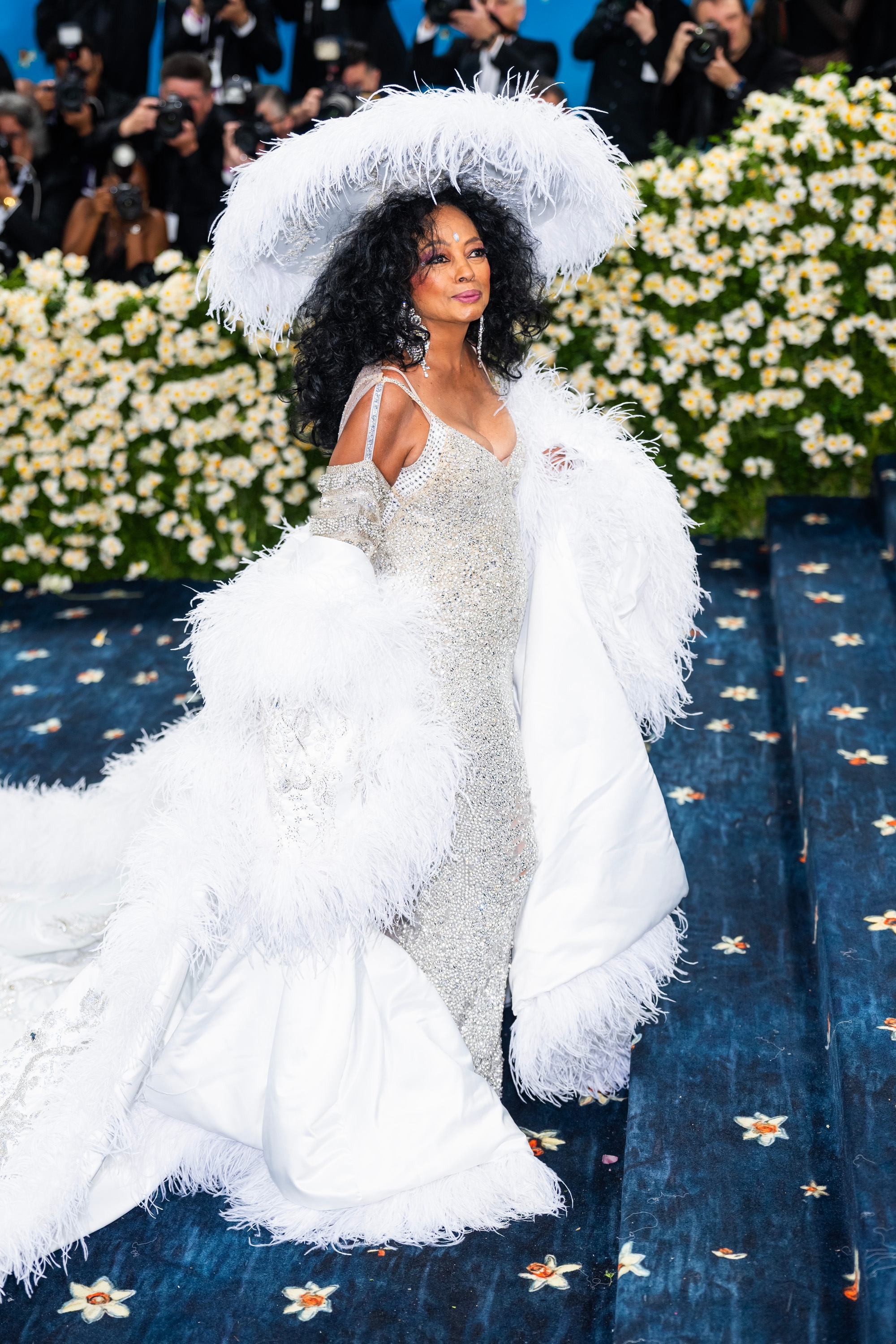 Person in a glamorous, feathered gown and hat on a starry blue staircase at a high-profile event