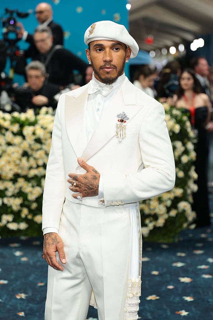 Individual in a white suit with a matching beret on a formal event carpet, surrounded by photographers and attendees in elegant attire