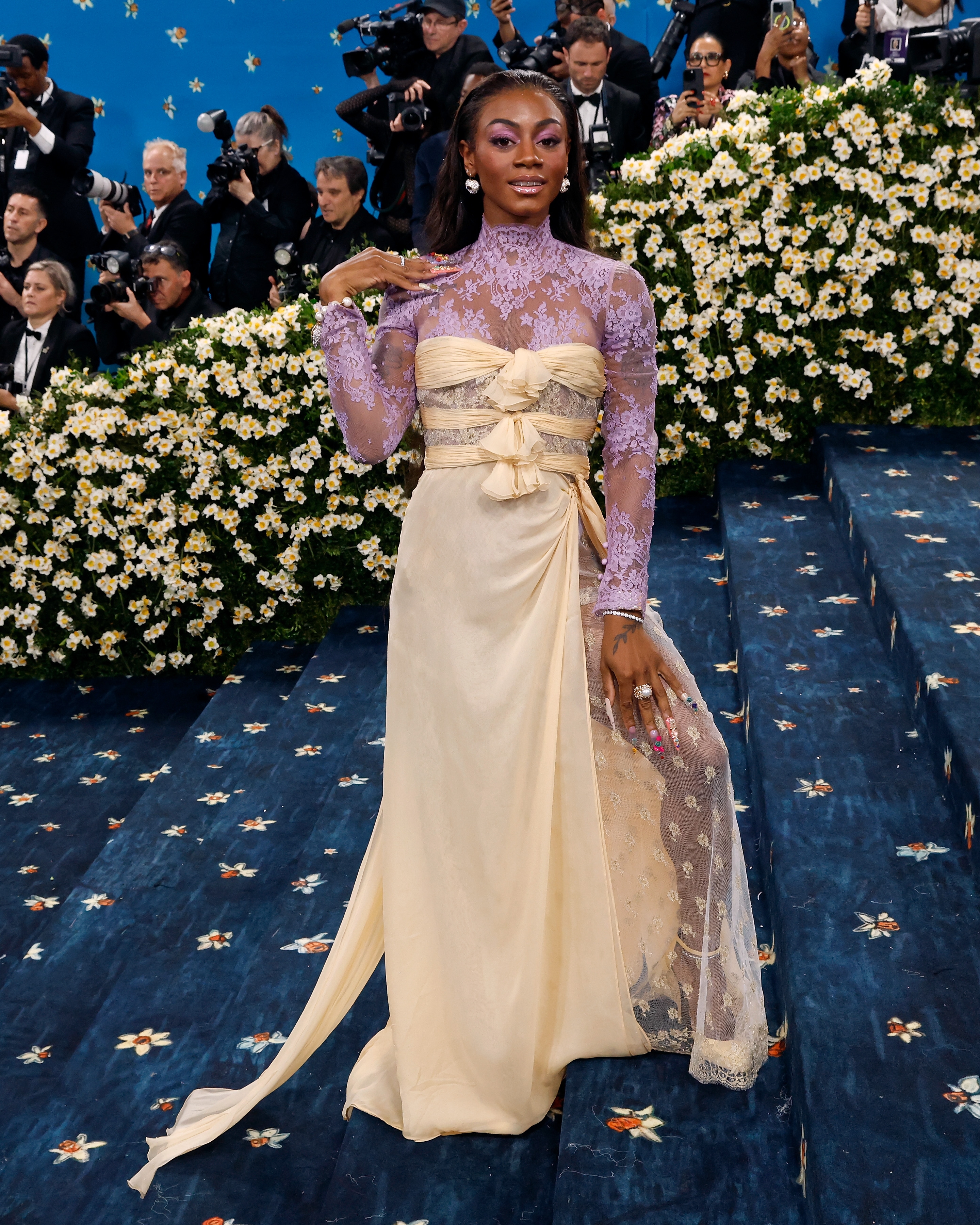 Person in elegant gown with intricate lace and ruffles poses confidently on floral-themed steps surrounded by photographers