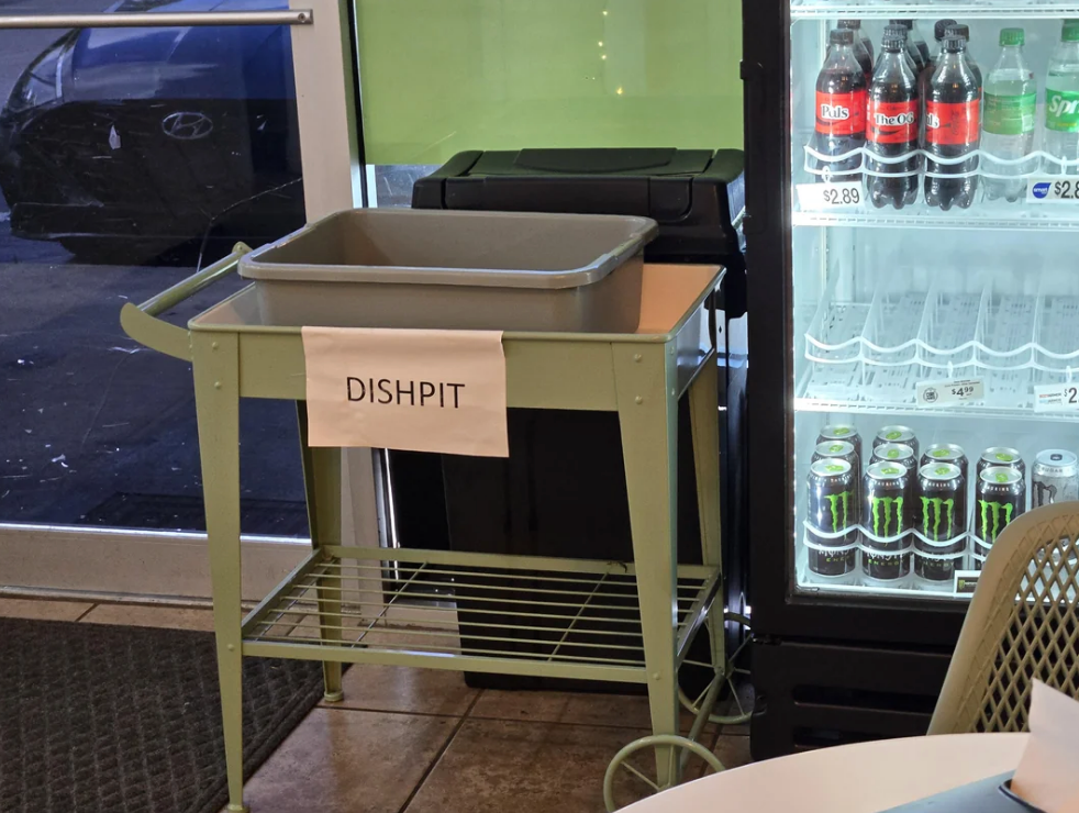 Dish cart labeled "DISHPIT" beside a fridge with drinks. At first glance, it appears to read "dipshit"