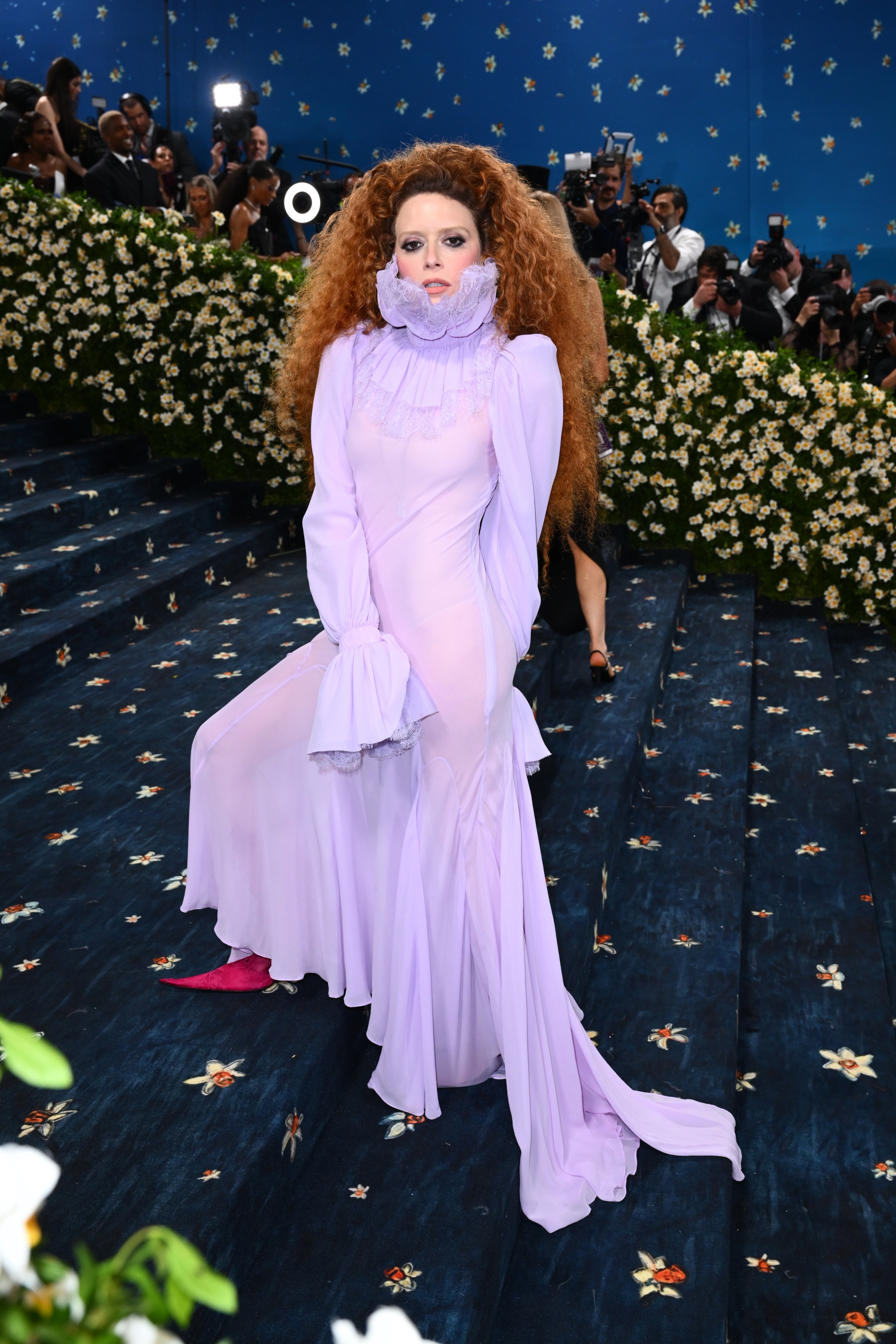 Person in a flowing, ruffled gown poses on a floral-adorned staircase amid photographers