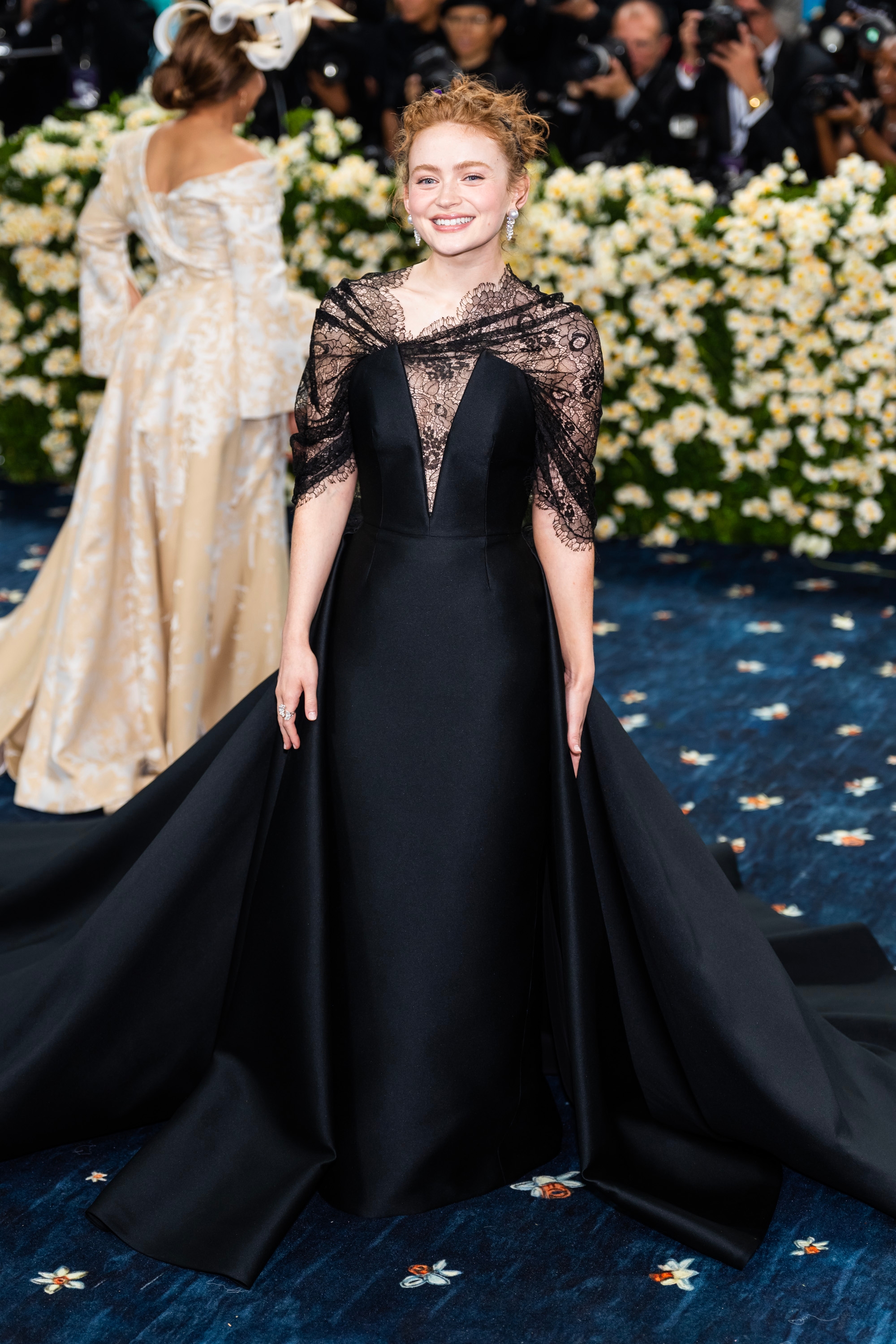 Celebrity on red carpet in an elegant black gown with a lace shawl and long train, smiling for photos with floral backdrop