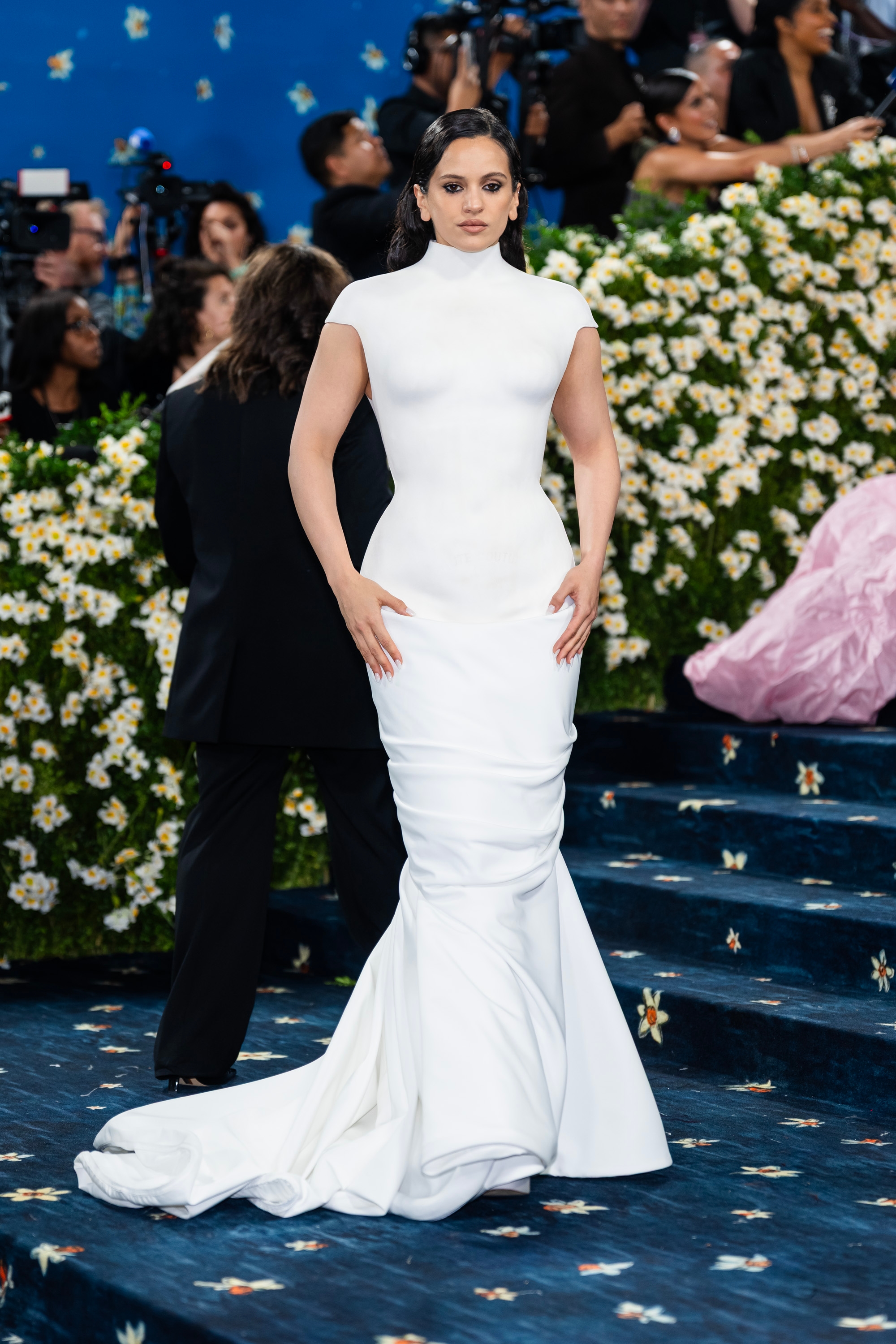 Rosalía in an elegant, form-fitting gown with a long train stands on a floral-decorated staircase at the Met Gala