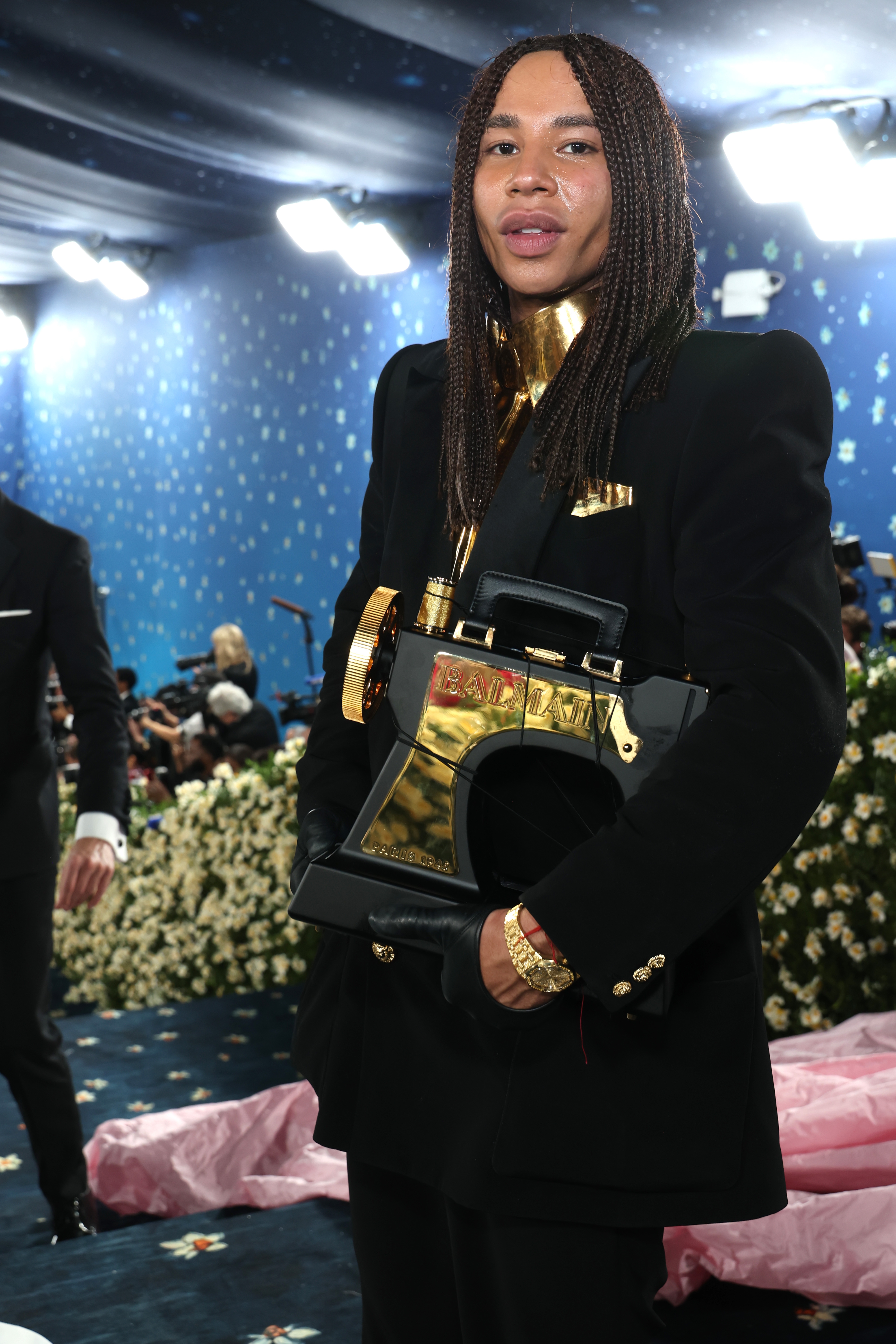 Olivier Rousteing on the Met Gala red carpet wearing a black suit with a gold collar, holding an oversized gold accessory