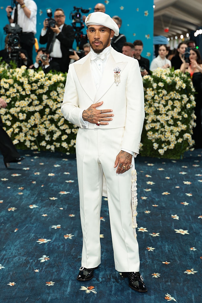 A man in an elegant, white suit with intricate details stands on a decorated carpet at a formal event, surrounded by photographers and attendees