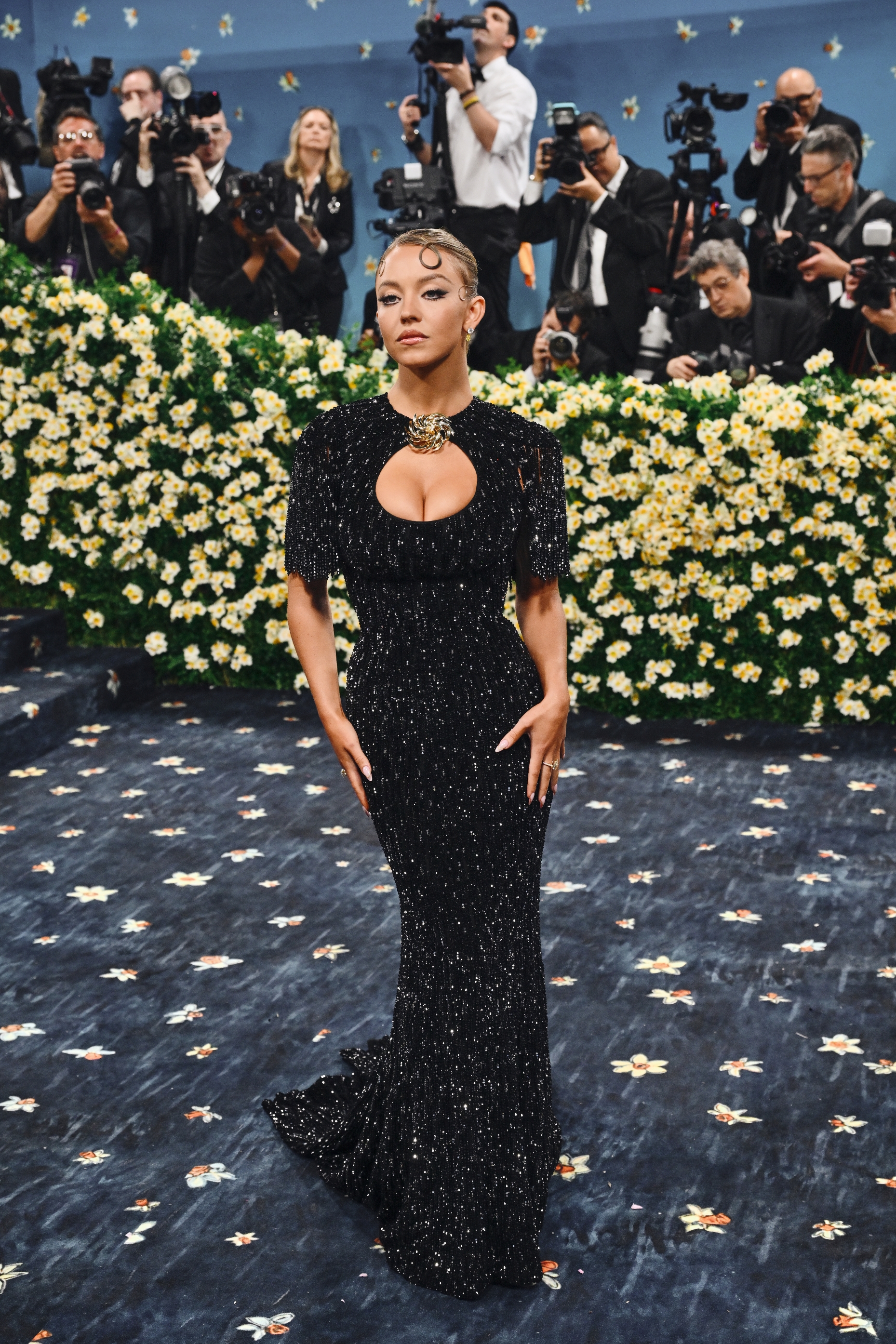 Person in an elegant, sparkling black gown with a keyhole neckline poses on a starry-themed carpet, surrounded by photographers