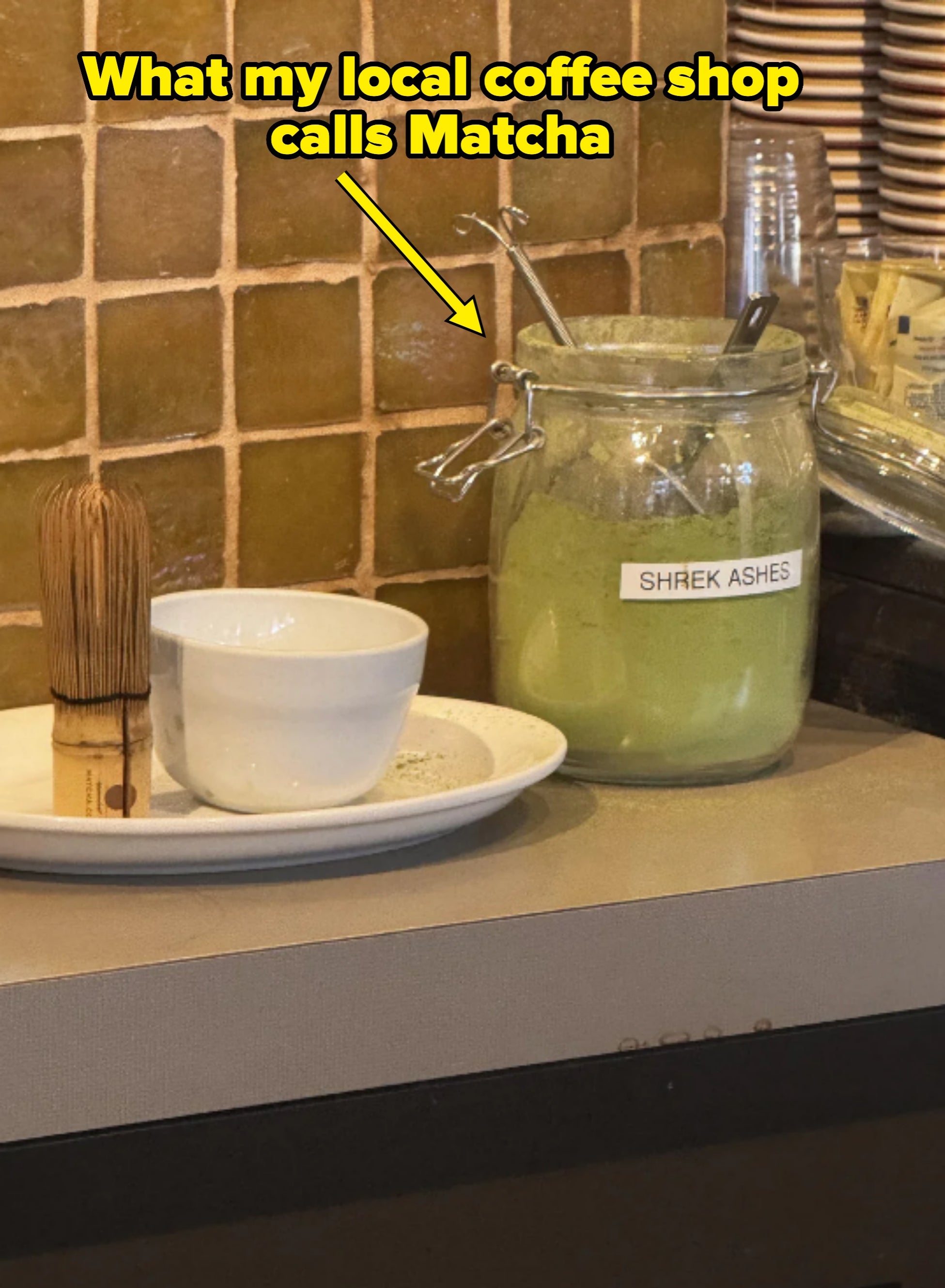 A jar labeled "Shrek Ashes" with green powder next to a bowl and matcha whisk on a counter. Caption: "What my local coffee shop calls Matcha"