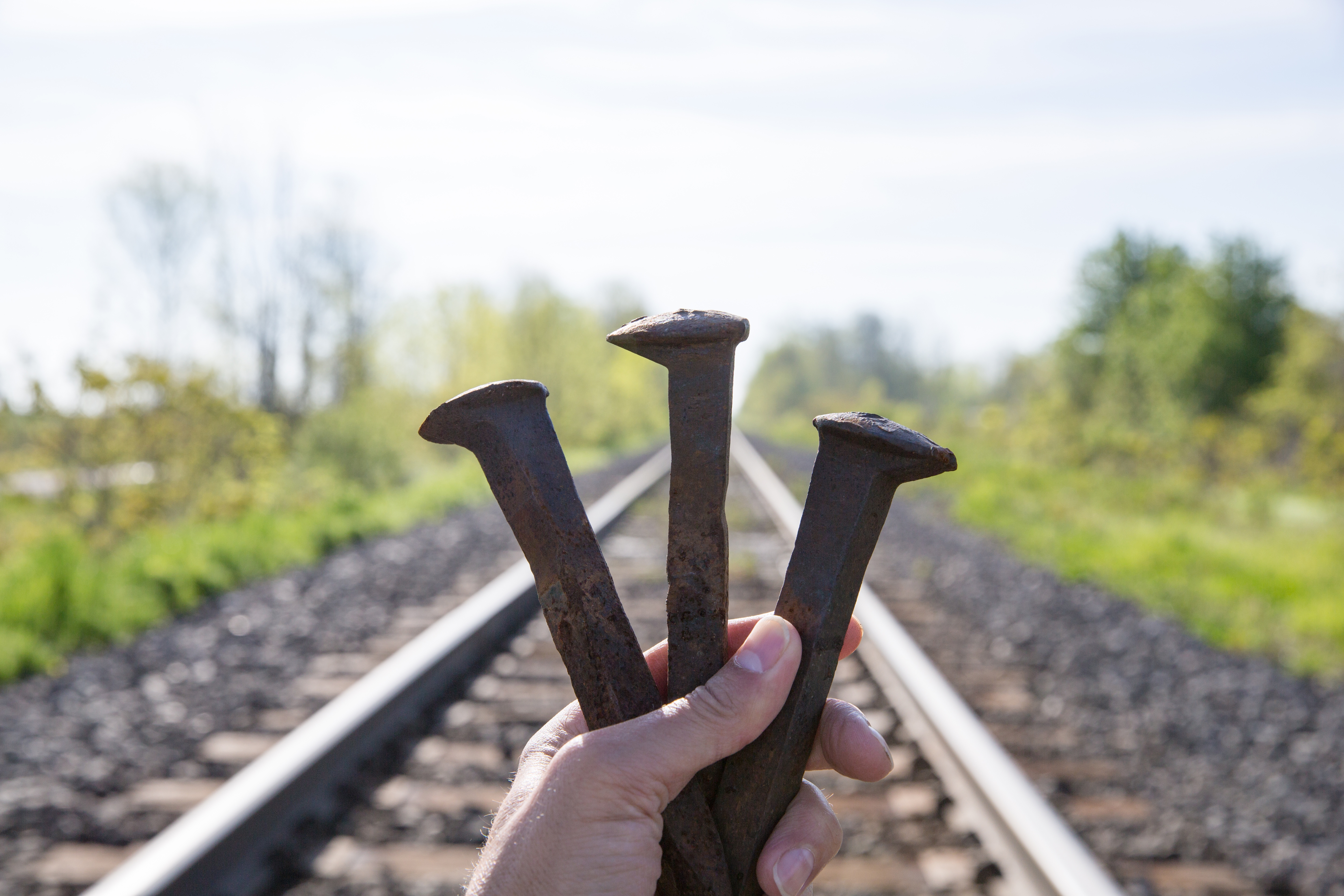 Hand holding three rusty railroad spikes against blurred train tracks background