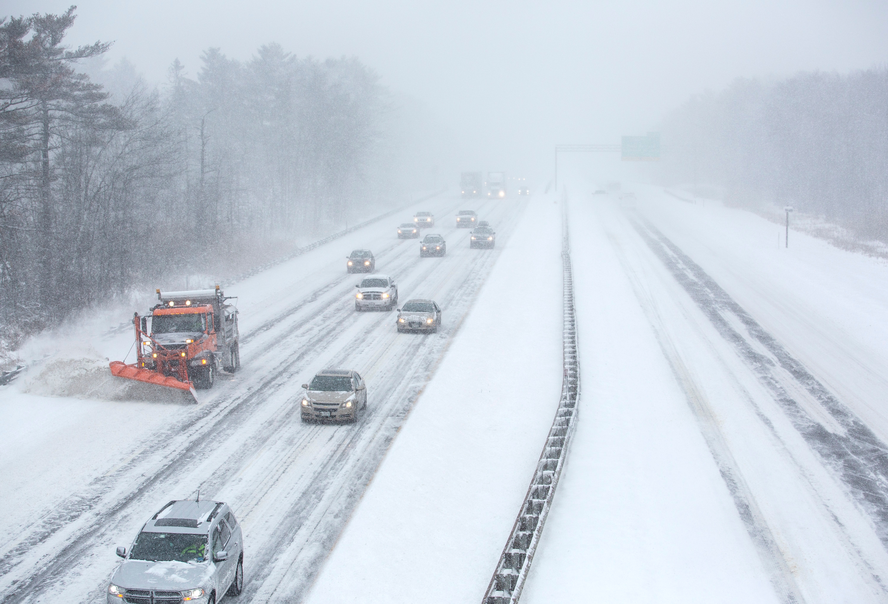 Cars and a snow plow drive on a snow-covered highway during a heavy snowfall, with limited visibility