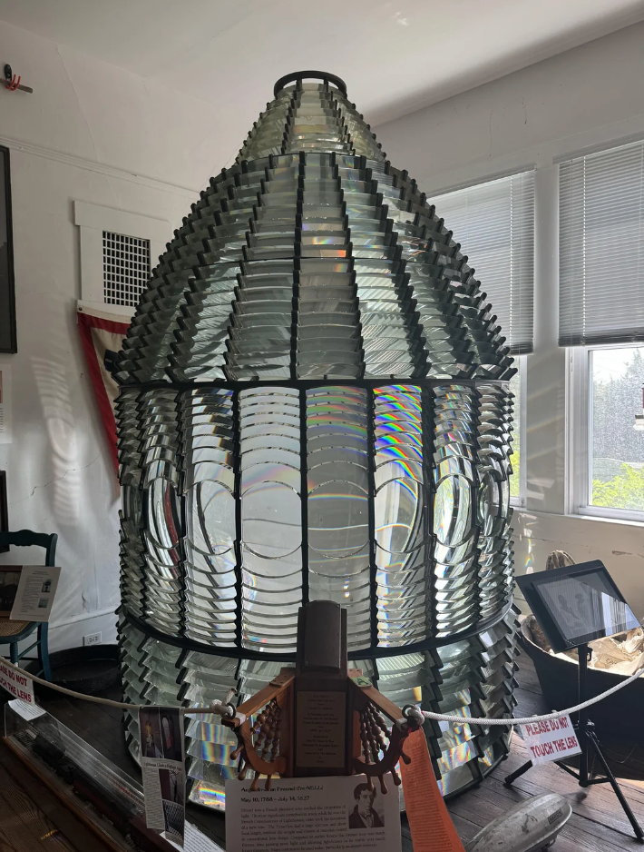 Large Fresnel lens display in a museum setting, with informational signs in the foreground and a window letting in natural light in the background