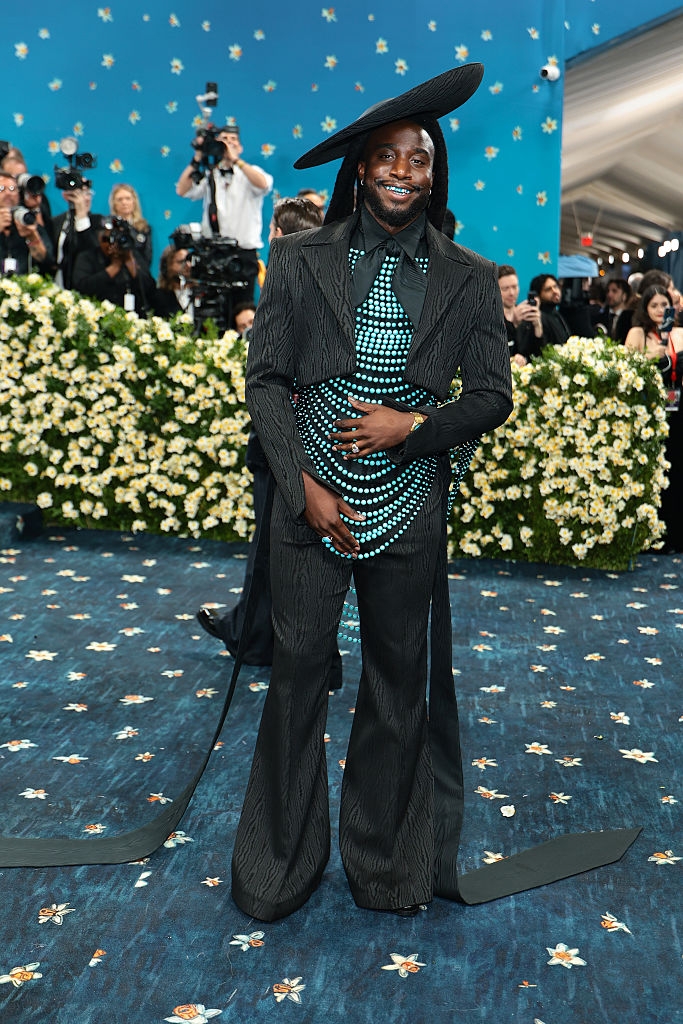 Person in a stylish outfit with a wide-brim hat and patterned top, posing confidently on a floral-themed event carpet