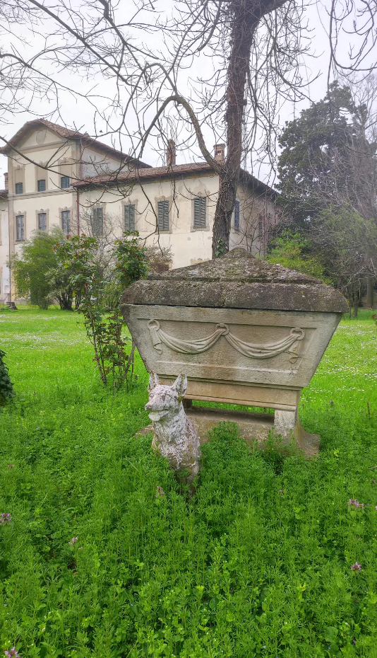 An ancient stone sarcophagus with draped fabric carvings stands on a grassy area in front of an old building with shuttered windows