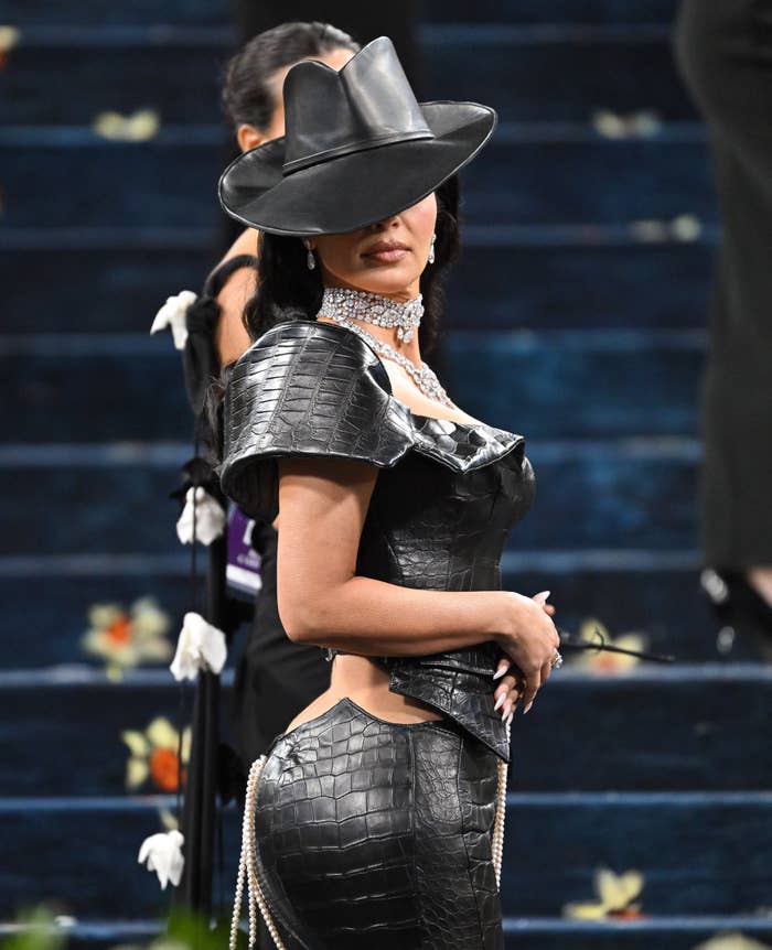 Person in avant-garde, off-shoulder leather dress with chain details and a wide-brim hat on a floral-patterned carpet, photographers in the background