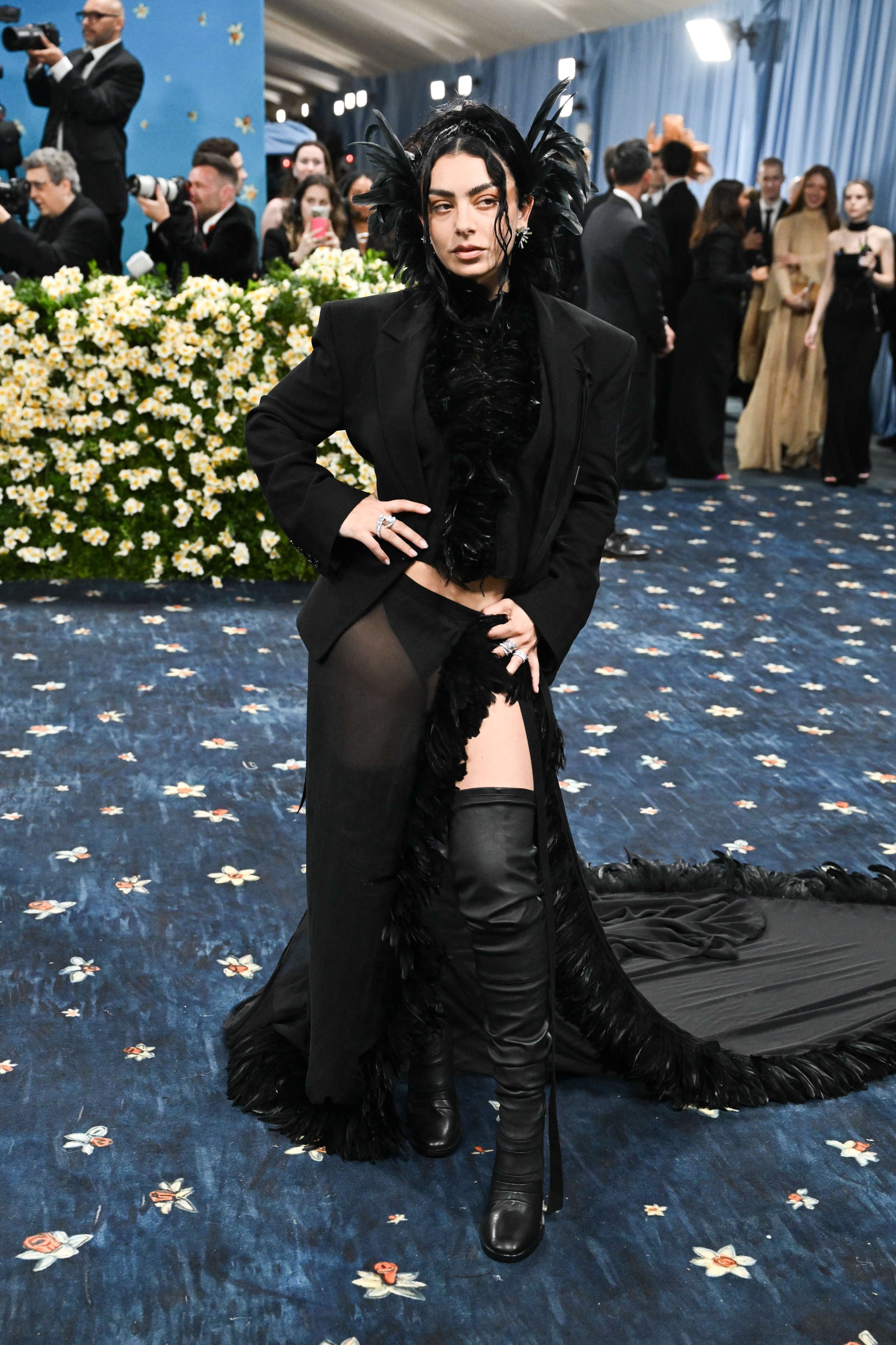 Person on a red carpet wearing a dramatic feathered and sheer black ensemble with a long train, striking a pose amid photographers and attendees