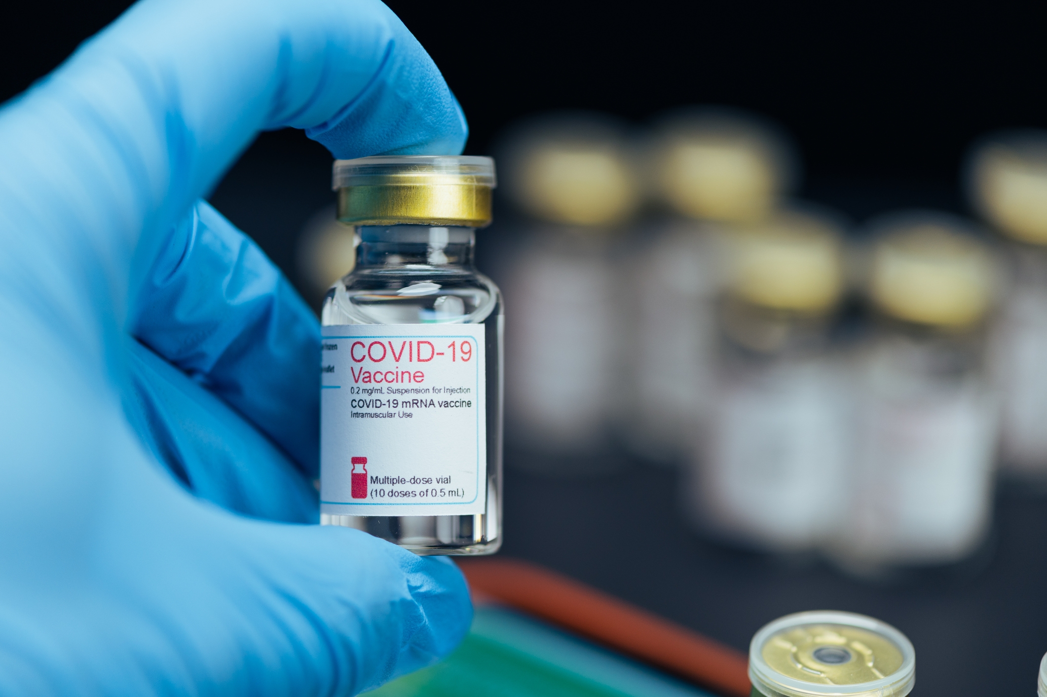 Gloved hand holding a COVID-19 vaccine vial, with more vials blurred in the background, signifying medical importance and safety