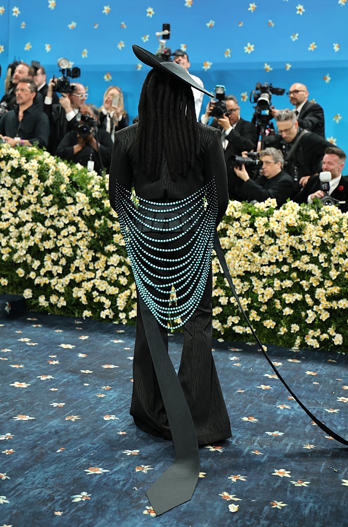 Person in avant-garde suit with pearl chains poses on a red carpet, surrounded by photographers