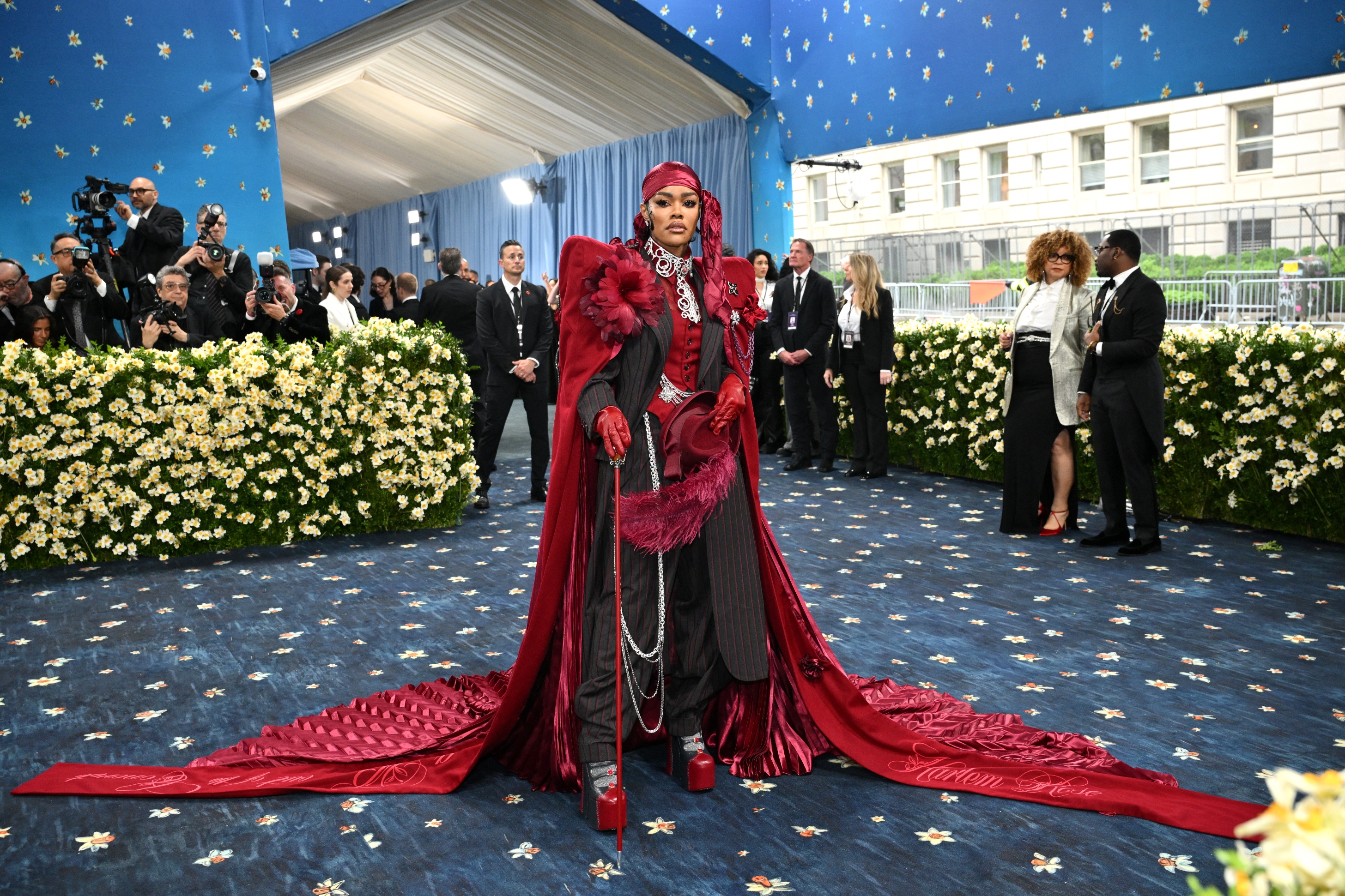 Person in elaborate, long train outfit with a cane at a formal event, surrounded by onlookers. The setting is upscale with floral decorations